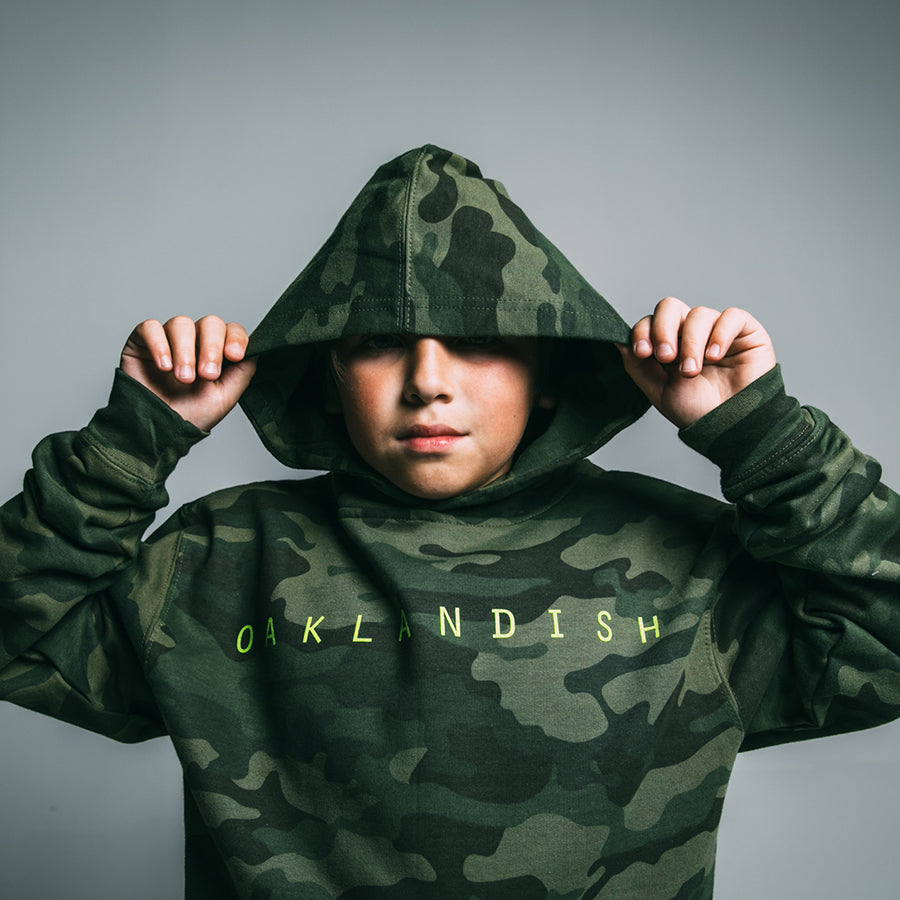 A youth sporting an Oaklandish Youth Classic Hoodie in green camouflage, featuring "OAKLANDISH" emblazoned in neon green letters on the front. With the hood pulled up over their head, they hold it with both hands against a solid grey background.
