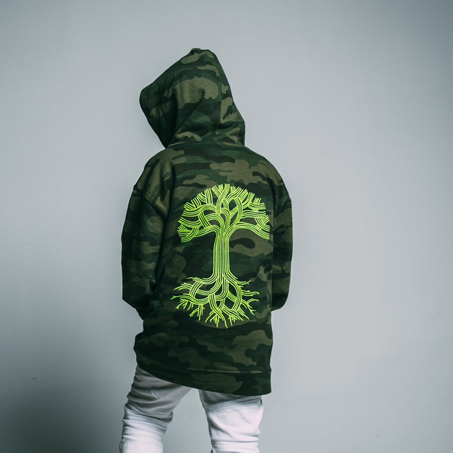 A person facing away from the camera against a plain light gray background is wearing an Oaklandish Youth Classic Hoodie in green camouflage with a neon green tree design on the back. The hood is up, covering their head, and they are paired with light-colored pants. This kids-sized hoodie is perfect for young adventurers.
