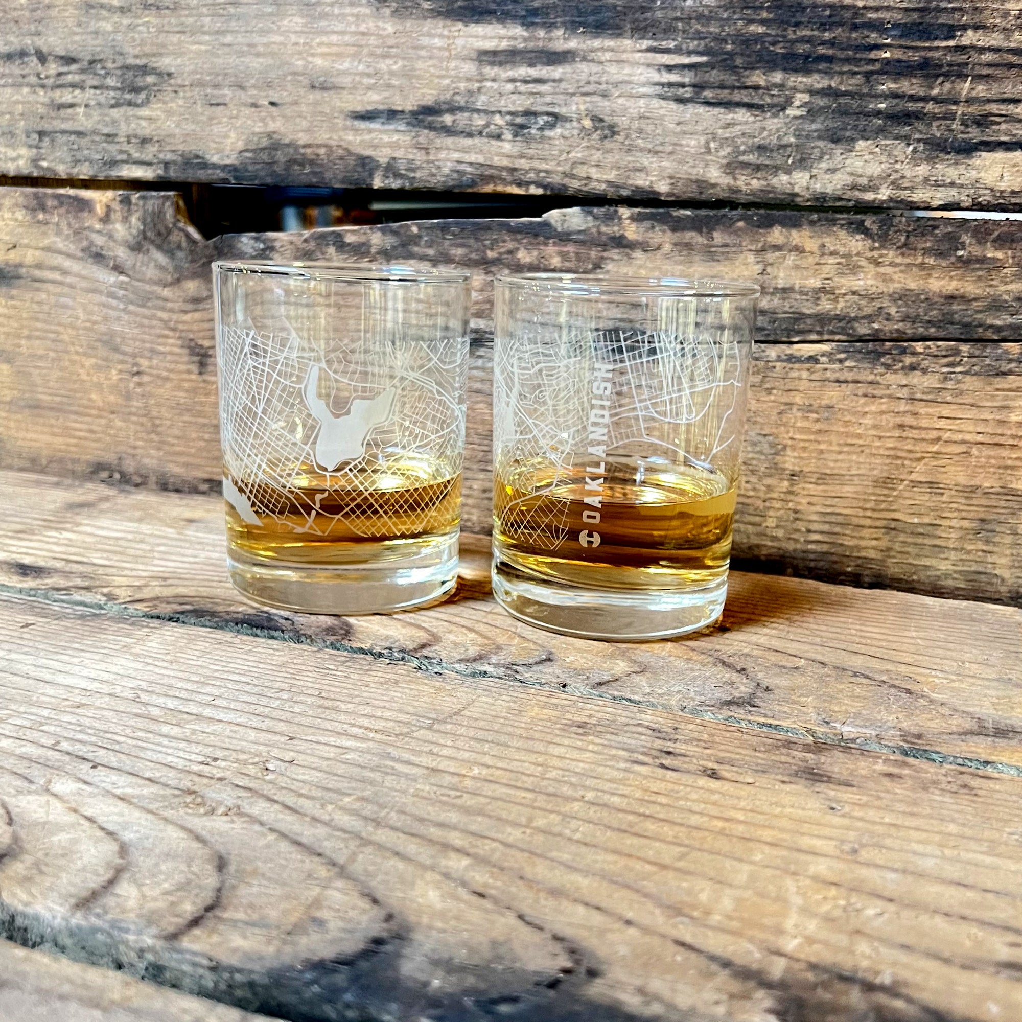 Two Aerial Tumblers by Oaklandish, each containing a small amount of amber-colored liquid, rest on a rustic wooden surface. Both classic glasses feature a white aerial map of Oakland etched onto them, adding a graphic element to the clear glass. The background consists of weathered wooden planks.