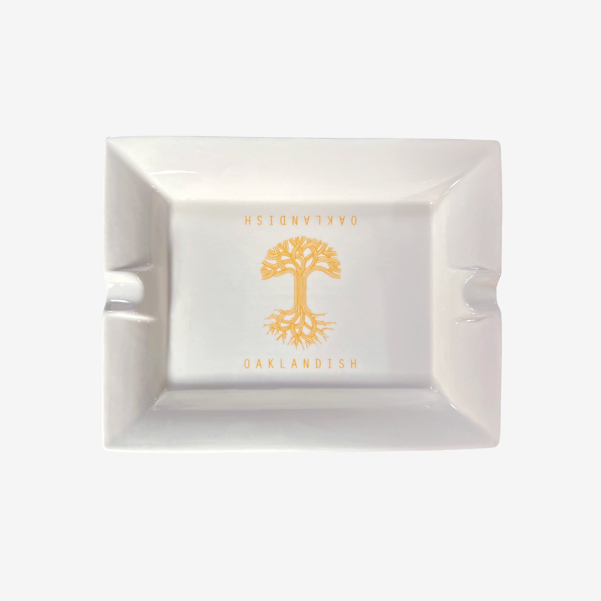The Oaklandish Ceramic Ashtray by Oaklandish is a rectangular, white ceramic ashtray with a simple design showcasing an orange tree with visible roots in the center. The "OAKLANDISH" logo is imprinted above and below the tree in orange capital letters. Additionally, two grooves on opposite sides of the ashtray are designed to hold cigarettes.