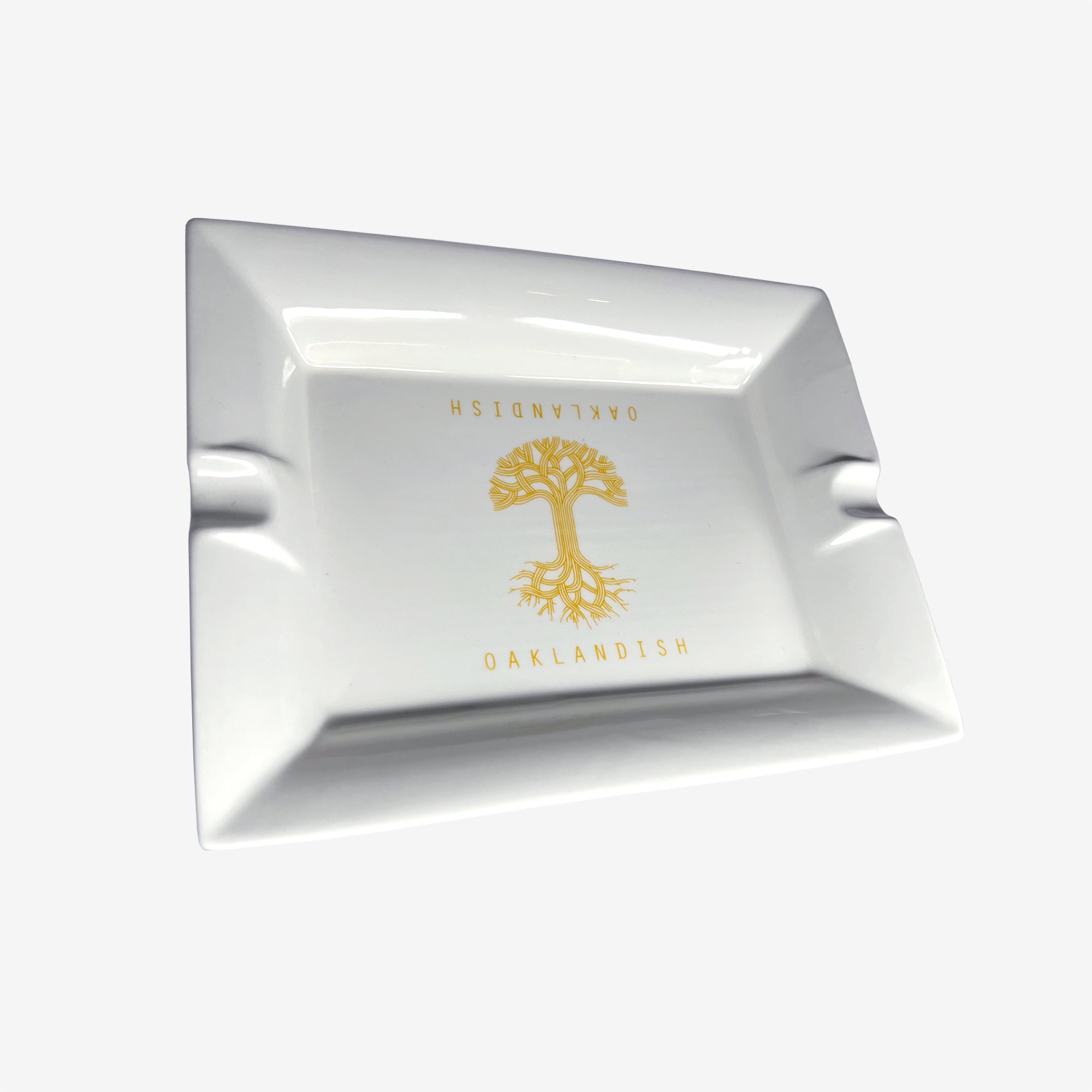 The Oaklandish Ceramic Ashtray, a rectangular white ceramic piece from the Oaklandish brand, is centered against a plain background. It features an imprinted Oaklandish logo and a gold tree emblem in the center. This ashtray boasts a glossy surface and has two indentations on opposite sides for resting cigarettes or cigars.