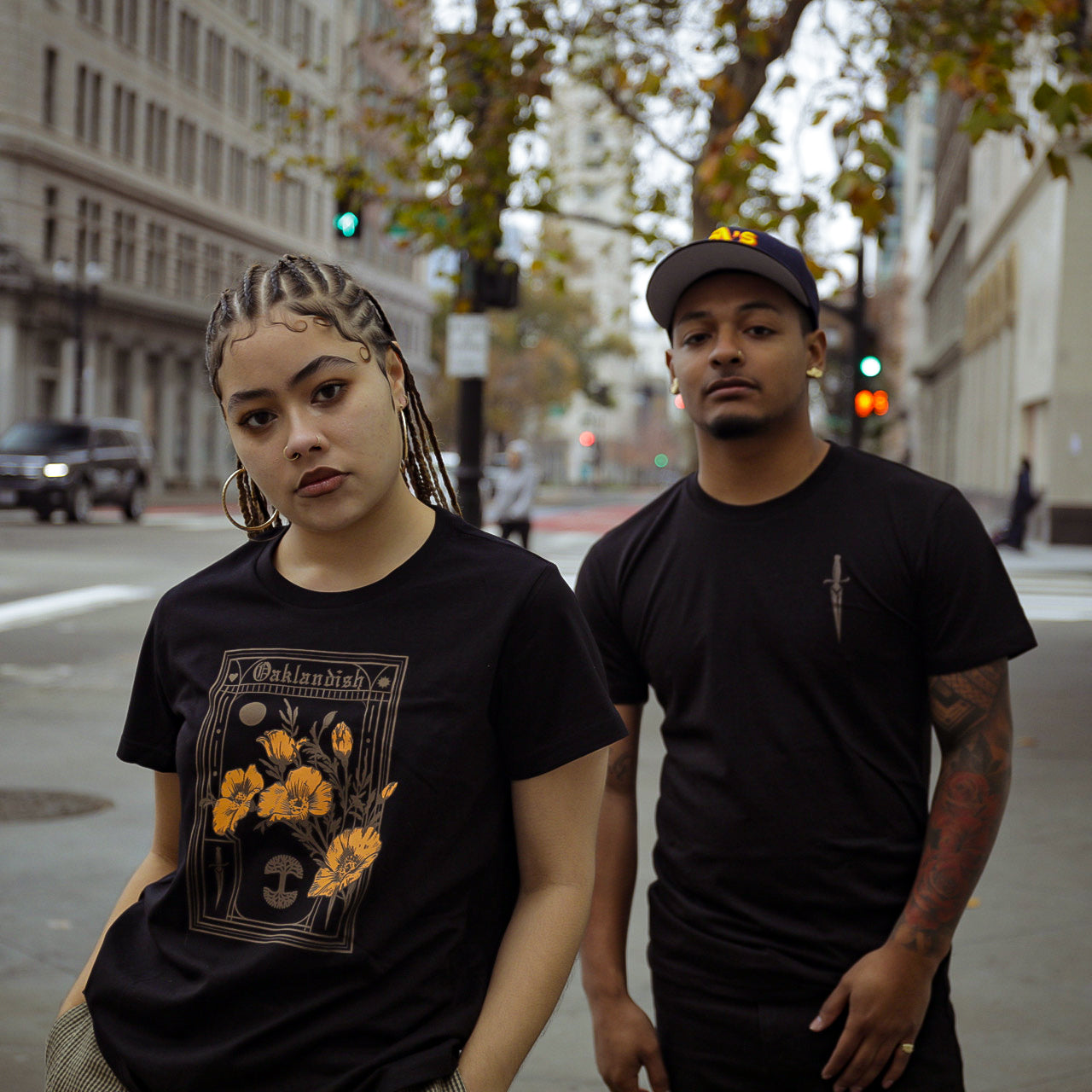 Two people stand in an urban street with buildings and trees behind them. The person on the left wears a black shirt with a floral and text design featuring poppies, has braided hair, and tilts their head slightly. The person on the right wears Oaklandish's Women's Blossom Tee, a cap, and has visible tattoos on their arms.
