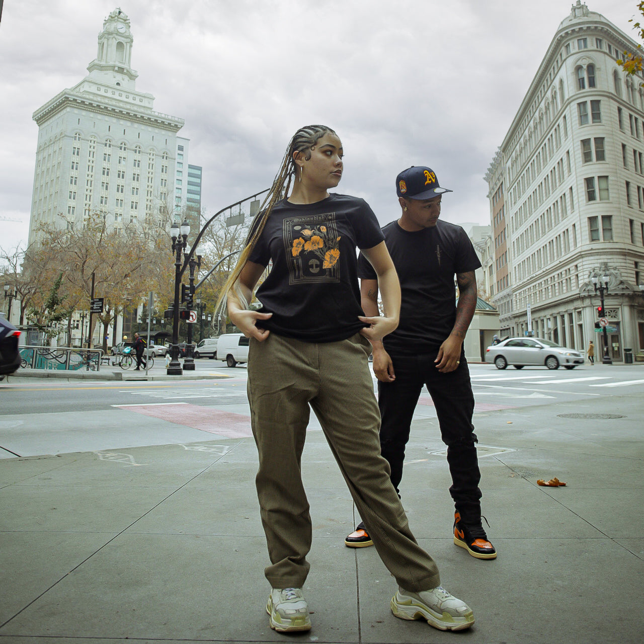 Two people stand on a city street. The person in the foreground, wearing an Oaklandish Women's Blossom Tee, gray pants, and white sneakers, has long braided hair. The person behind wears a black baseball cap, a black T-shirt in women's sizing, and black pants with orange and black sneakers. Buildings and trees are in the background.