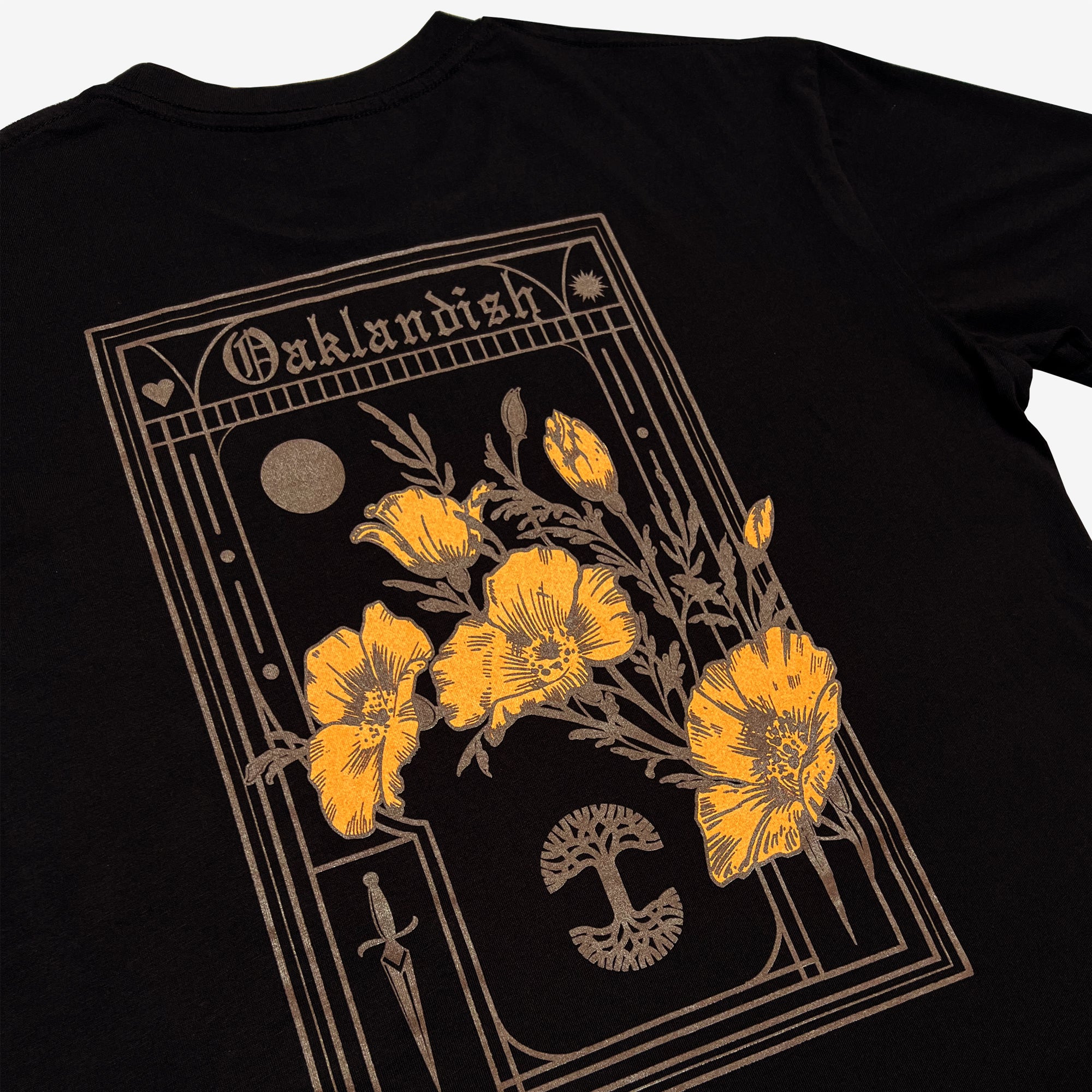 Oaklandish's Blossom Tee is a black men's cotton t-shirt with an intricate design. The design features a large rectangle at the top with the word "Oaklandish", a moon, a heart, and a dagger. Inside the rectangle are several yellow poppy flowers—the California state flower—displayed with detailed stems and leaves over a black mushroom graphic.