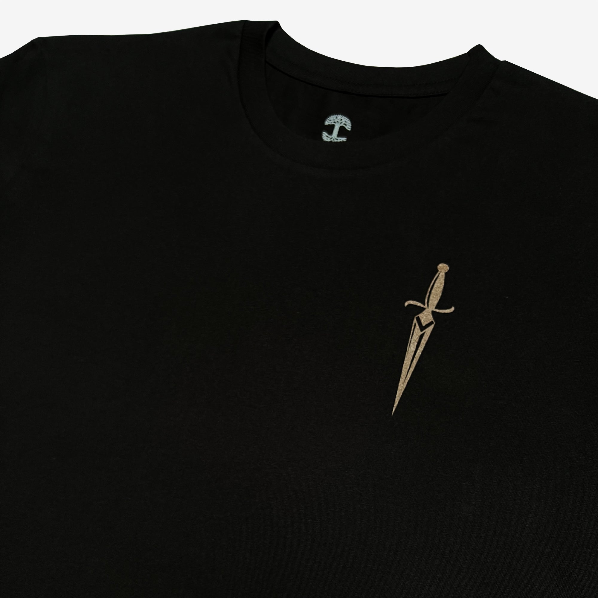 A close-up of the Oaklandish Blossom Tee, a black men's cotton T-shirt featuring a small, intricately designed gold dagger emblem on the upper left chest. The dagger has a sharp, pointed blade and an elegant hilt with stylized details. The plain white background makes the T-shirt and dagger design stand out beautifully.