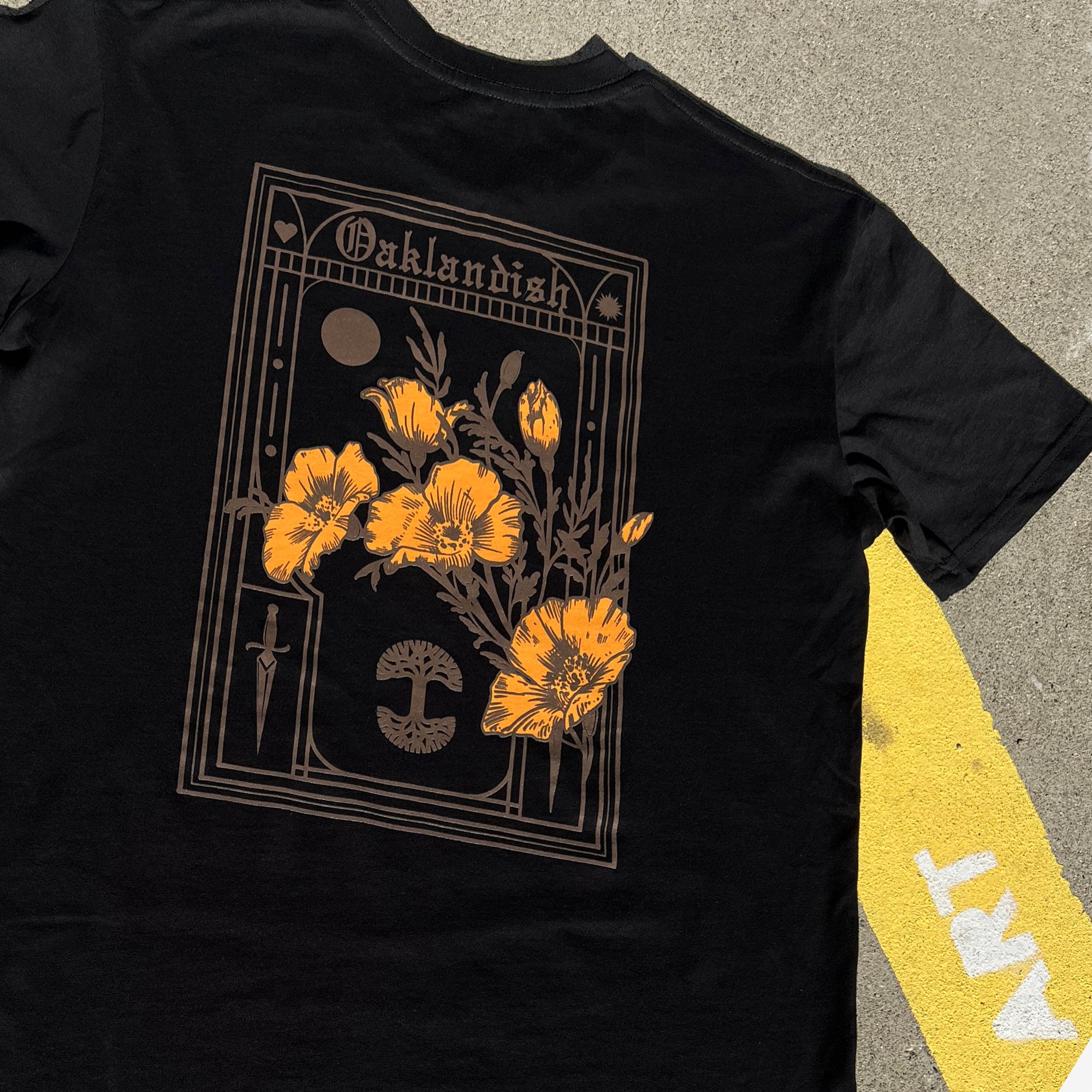 The Blossom Tee by Oaklandish is a black men's cotton t-shirt adorned with an elaborate back design. It features the word "Oaklandish" at the top, surrounded by yellow-orange poppies, a dagger, a full moon, and other detailed elements that frame the central floral motif. The shirt is displayed on a concrete surface with a yellow and white striped background.