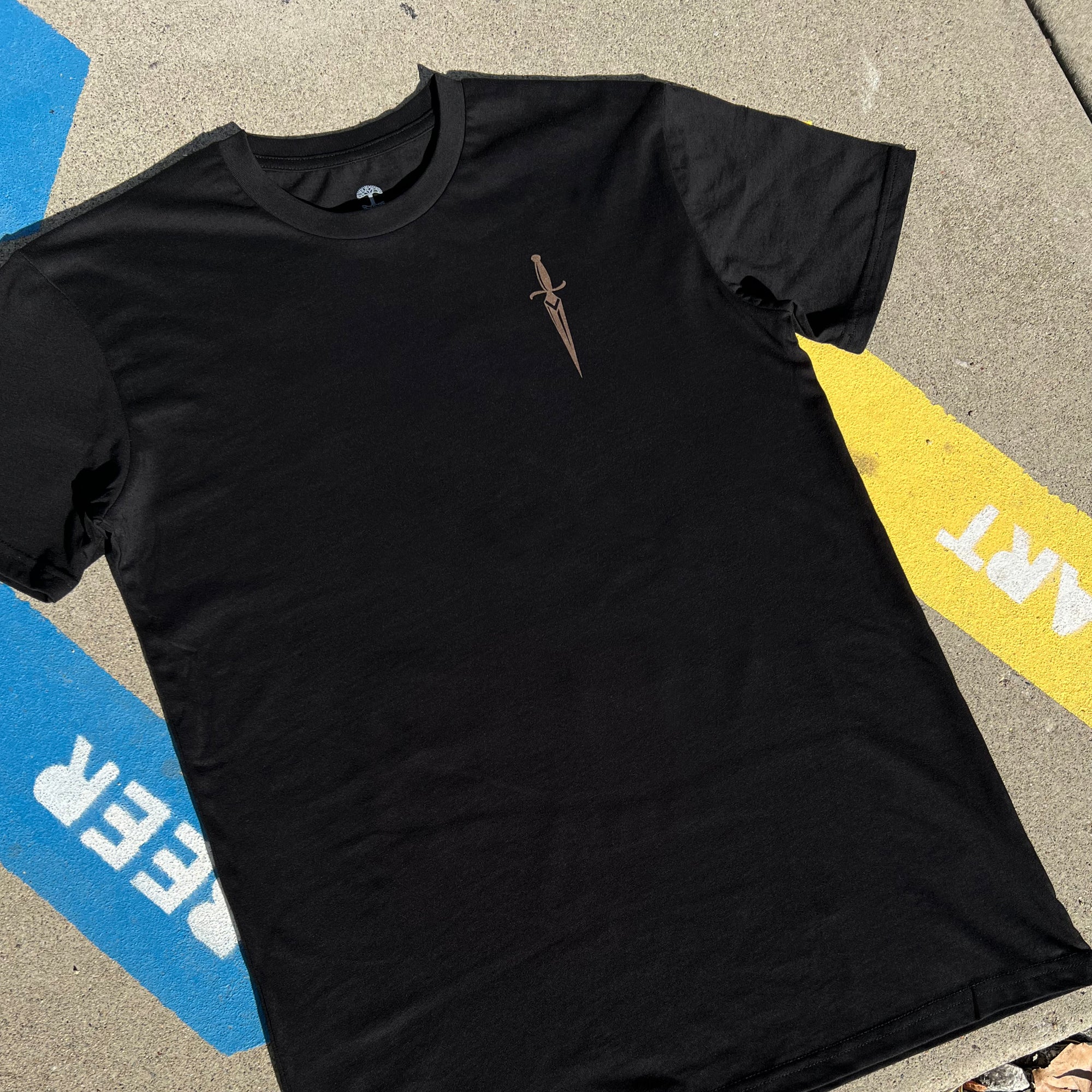 The Blossom Tee by Oaklandish, a black men's cotton t-shirt, is laid flat on the ground. It features a small brown dagger graphic on the left chest area. In the background, concrete with blue and yellow painted markings displays partially visible words "REER" in blue and "ART" in yellow, adding character to the scene.