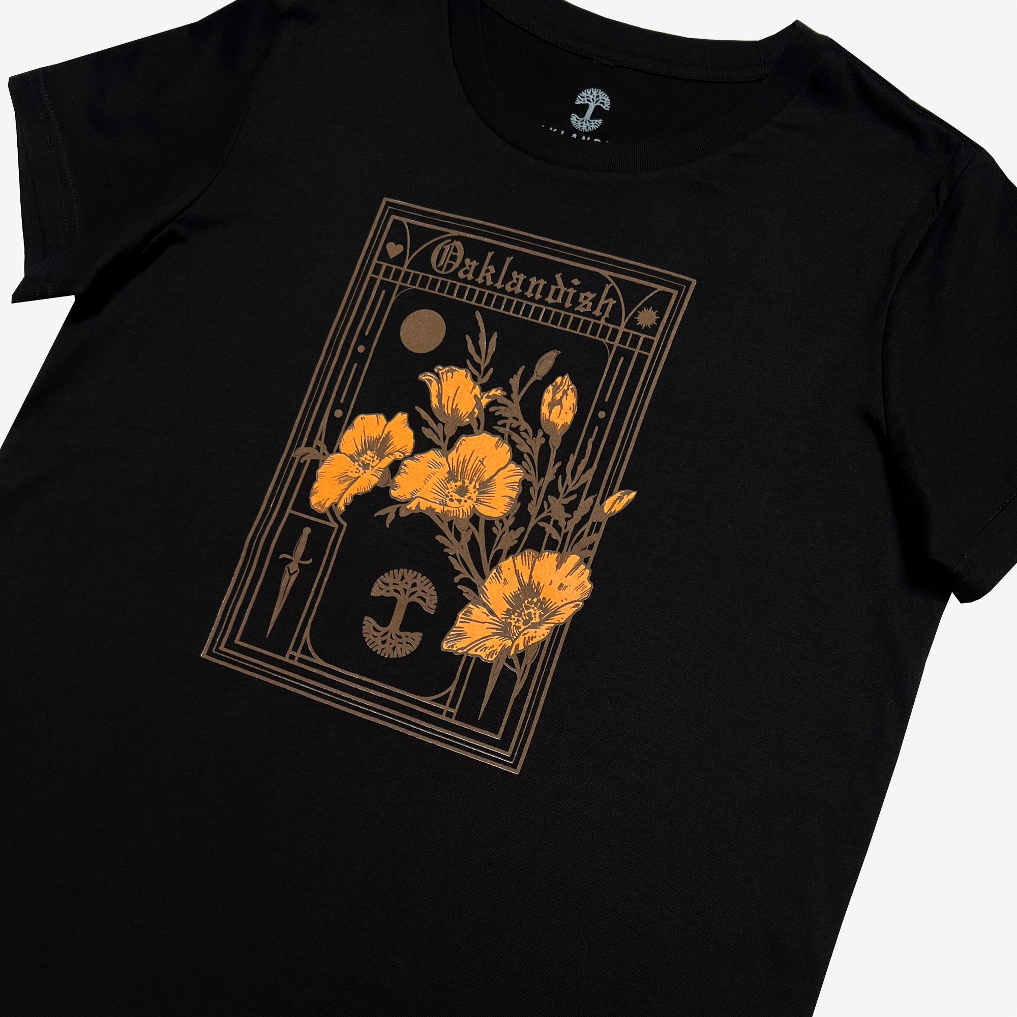 The Women's Blossom Tee by Oaklandish is a black t-shirt featuring a stunning golden floral design on the front. The intricate design showcases a bouquet of poppies framed by an ornate border, along with geometric elements like a crescent moon, a dagger, and a circular motif. "Oaklandish" is written in a stylized font at the top and it is available in women's sizes.