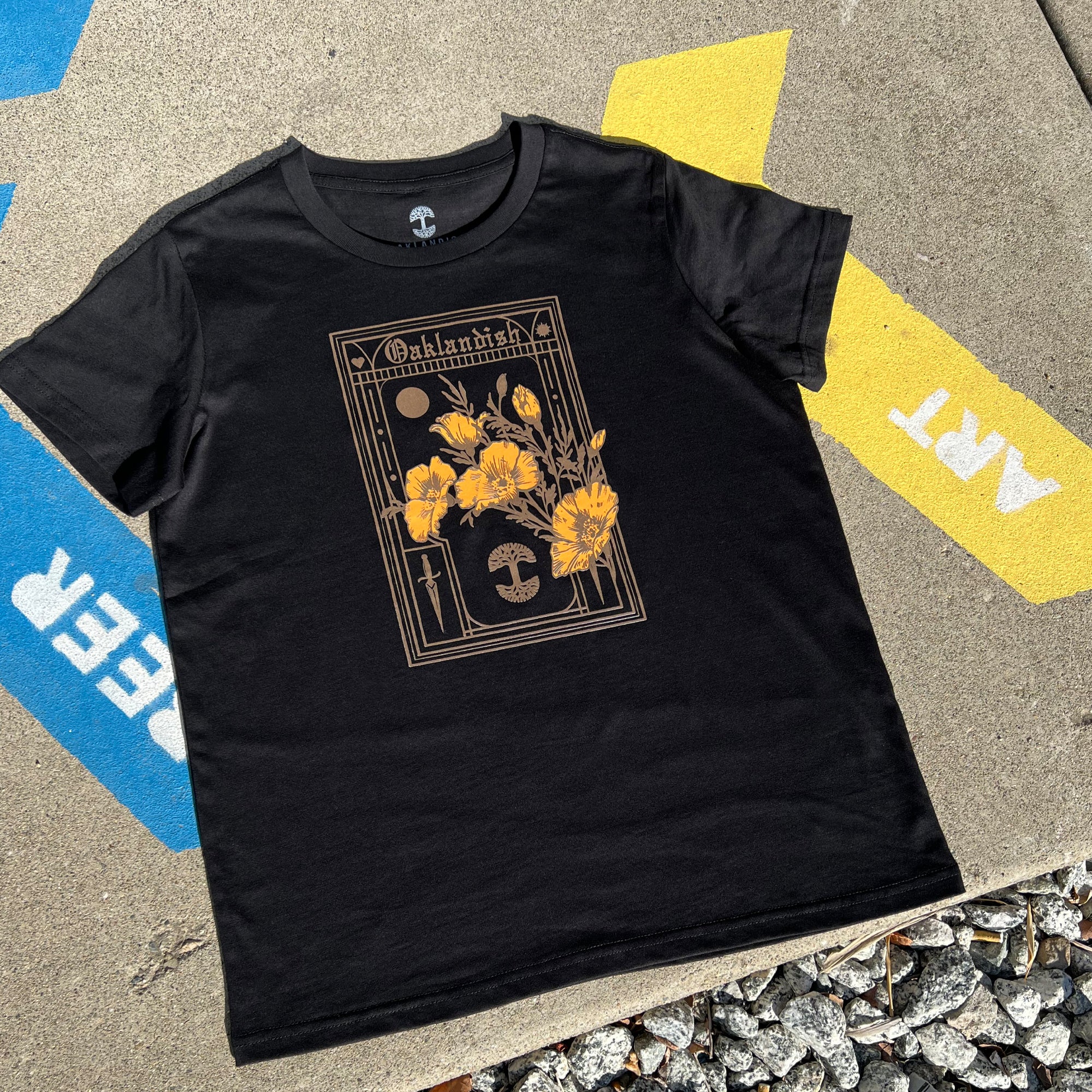 The Women's Blossom Tee by Oaklandish is a black T-shirt in women's sizing that showcases a detailed design on the front with yellow poppies, a moon, and the word "Oklahoma" framed ornately. The shirt is photographed laid flat on concrete pavement, partially over a yellow and blue painted section featuring the white text "FIRE" and "ART," with small rocks scattered at the bottom of the image.