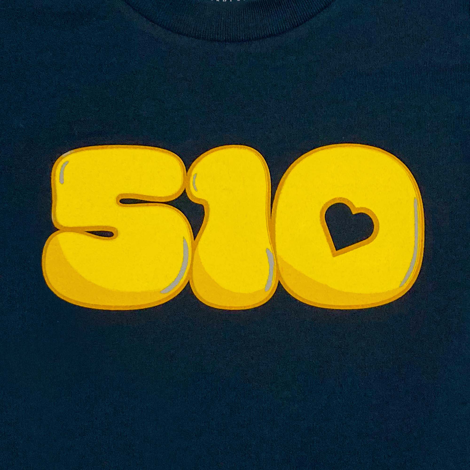 The Toddler Bubble 510 Tee by Oaklandish features a bold yellow graphic design with inflated numbers "510" on a navy, toddler-fit t-shirt. A small heart shape cutout in the upper right corner of the "0" adds a playful touch to the overall design, and subtle highlights give the numbers a 3D effect.