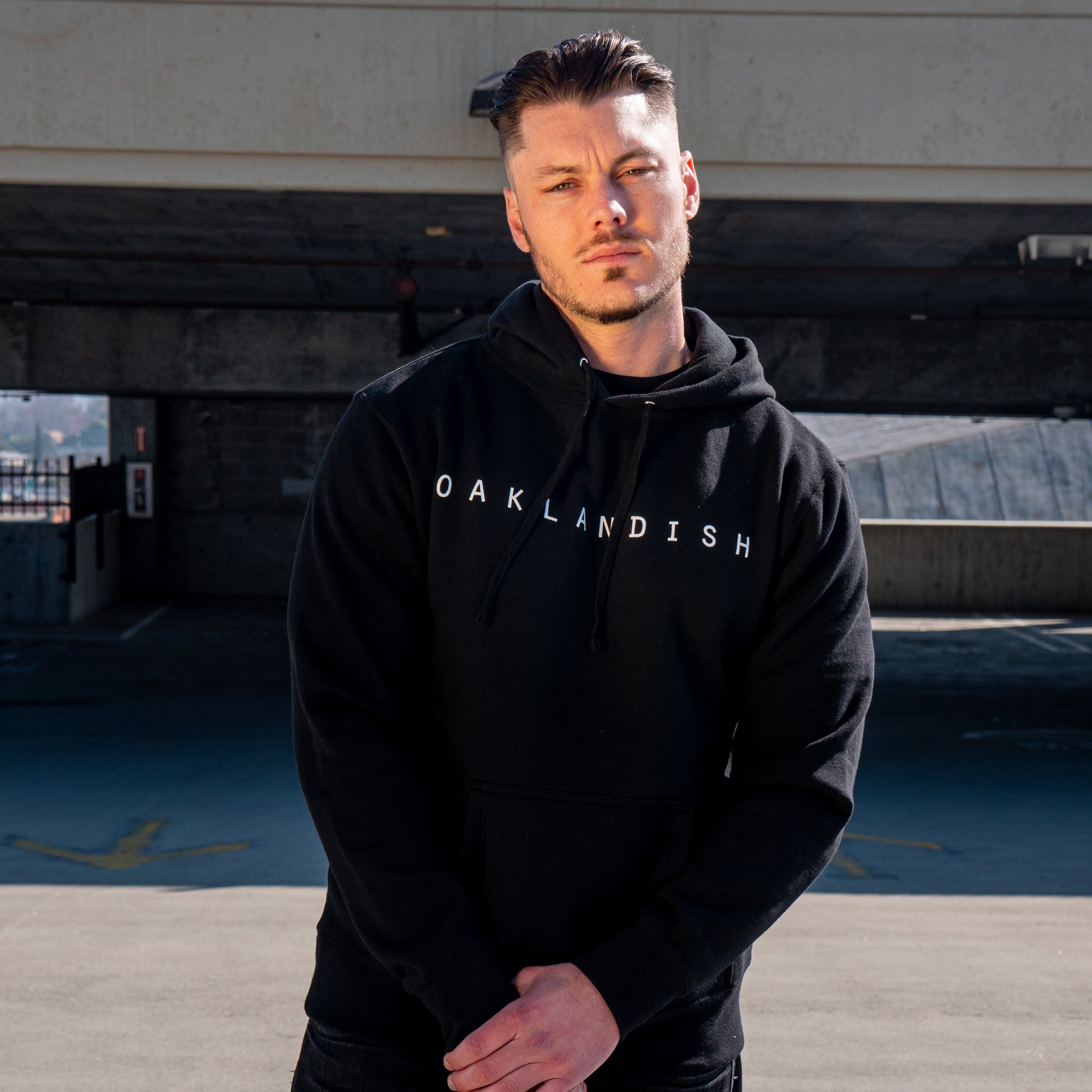 A man with short, styled hair and a trimmed beard stands outdoors, wearing an Oaklandish Classic Hoodie from the brand Oaklandish. The black pullover hoodie prominently features the word "OAKLANDISH" in white letters. Behind him is a concrete structure with partially shaded areas and hills visible in the distance. His serious expression reflects his Oakland roots as he gazes directly into the camera.