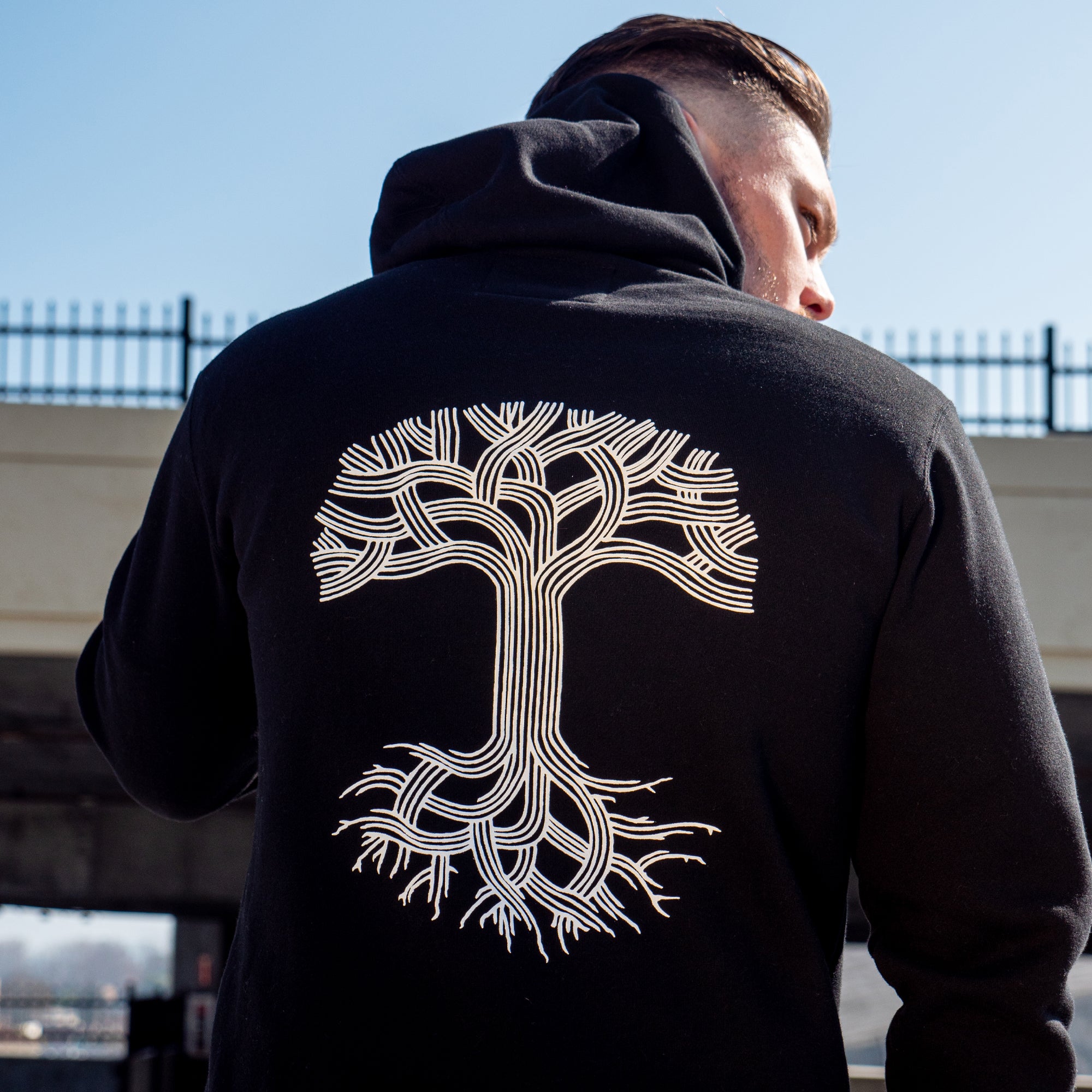 A person with short hair is wearing an Oaklandish Classic Hoodie, a black pullover featuring a large, stylized white tree graphic on the back that evokes Oakland roots with its intricate, interwoven branches and roots. The person is standing outdoors near a metal fence and an underpass.