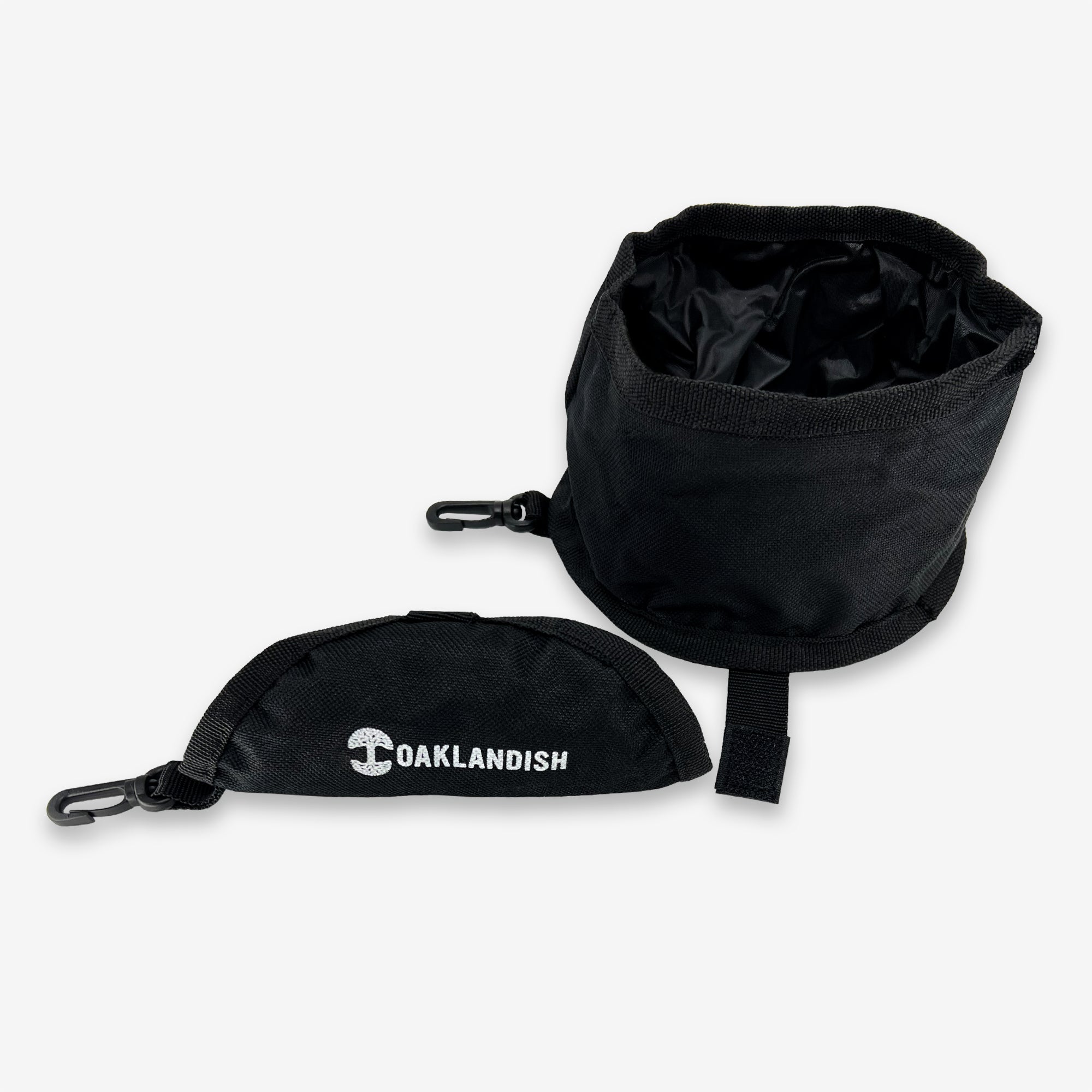 The Travel Pet Bowl by Oaklandish is a black collapsible pet bowl made from durable 600D polyester, featuring a smooth inner lining and a small loop for attaching the included carabiner. Displayed in both its expanded and folded states, the folded version proudly bears the "OAKLANDISH" print in white, making it an ideal portable accessory for your pet.