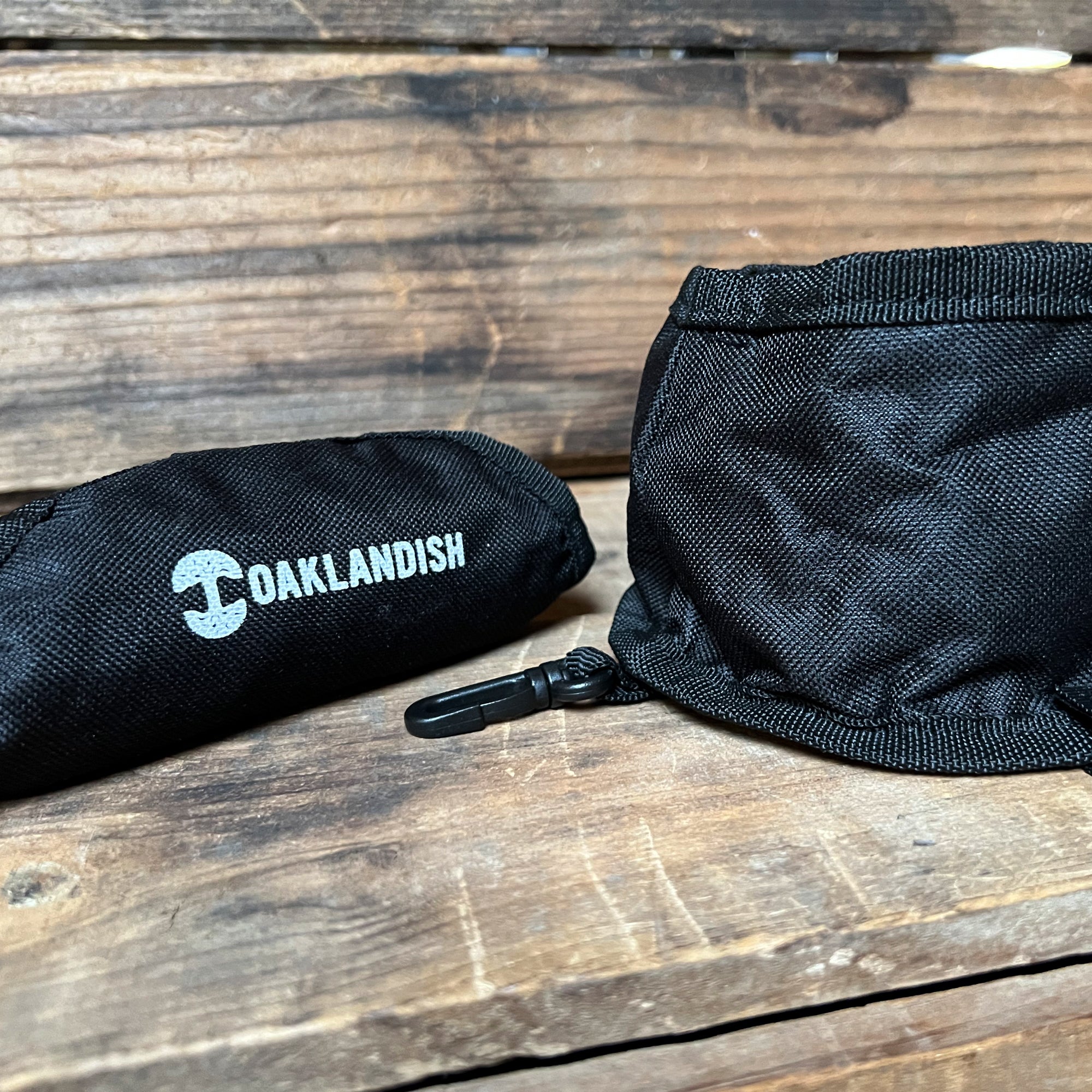 A small, rolled black Travel Pet Bowl made of durable 600D polyester with the word "Oaklandish" printed in white rests on a rustic wooden surface. Next to it lies an unfolded section of a similar black fabric pouch, revealing a zipper and clip, hinting at its use as a portable pet accessory.