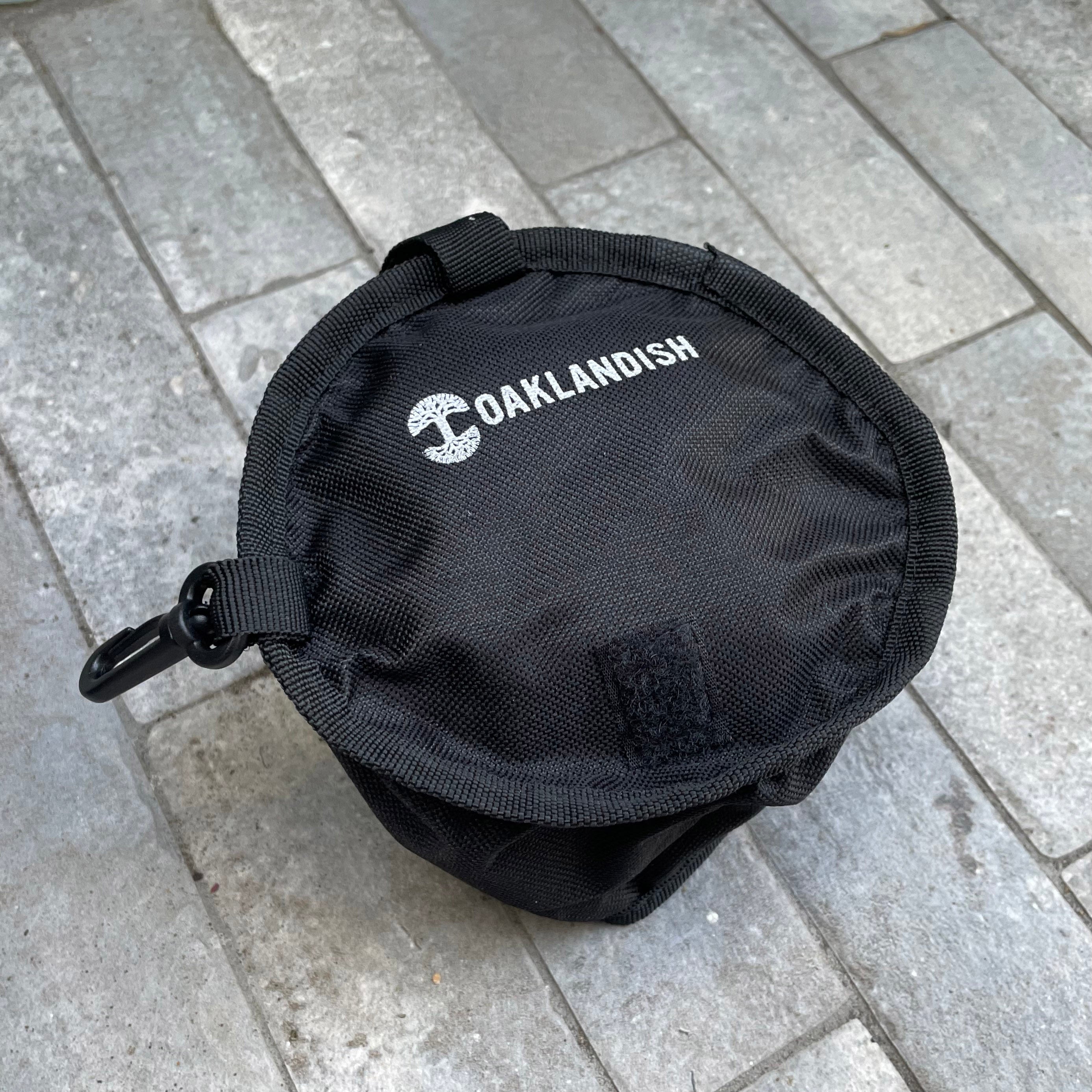 A compact black pouch featuring a zipper and a clip on one side rests on a grey brick pavement. On the top of the pouch, "Oaklandish" is printed in white along with their distinctive tree logo. Crafted from robust 600D polyester, it's ideal for outdoor or utility purposes, possibly for storing an Oaklandish Travel Pet Bowl.