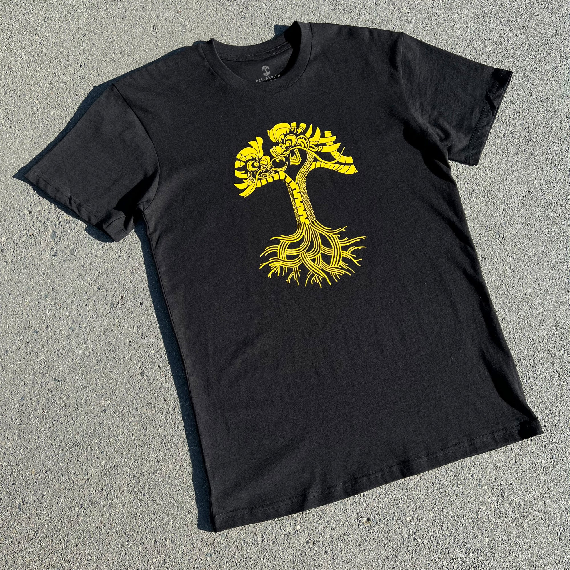 Image of the Dragon Power Tee by Oaklandish laid on a flat surface. The black T-shirt showcases a striking yellow graphic design on the front, featuring a stylized tree with intricate, swirling roots and branches reminiscent of dragons. The vibrant design stands out against the dark fabric.
