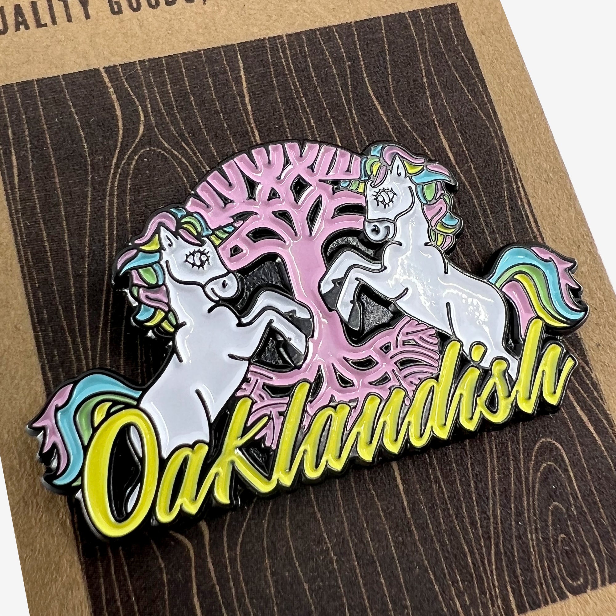 Introducing the Elusive Enamel Pin by Oaklandish: This beautifully designed pin features two white unicorns with multicolored rainbow manes, rearing up beside a pink tree with dark brown branches. The word "Oaklandish" is presented in yellow and outlined in black below the unicorns. The pin comes attached to a textured brown backing card.