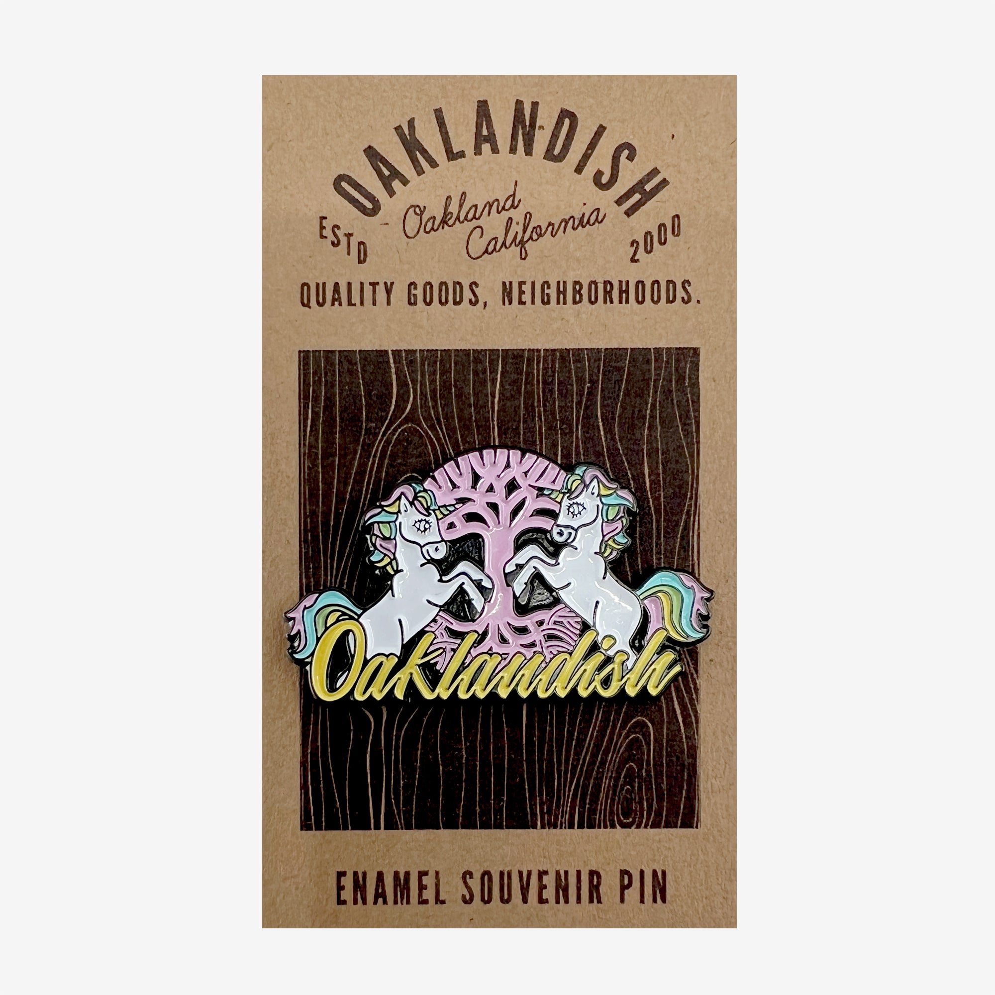 The Elusive enamel lapel pin by Oaklandish showcases two white unicorns with multicolored manes flanking a stylized pink tree, adorned with "Oaklandish" in yellow cursive. The background has a wooden texture, and the card features "Oaklandish" and "Oakland, California." It's the perfect addition for accessories lovers!