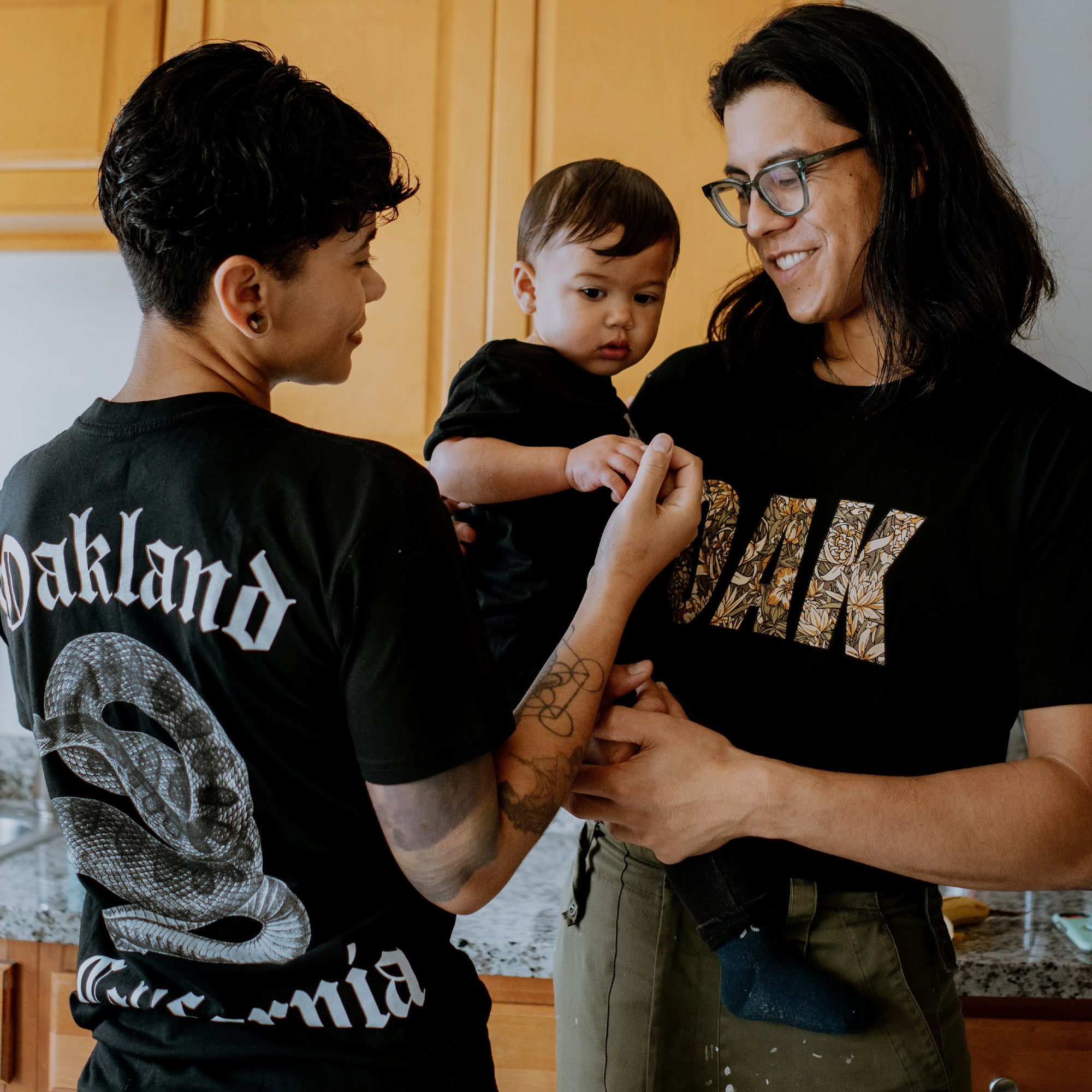 Two adults are holding a baby in a kitchen, all wearing black T-shirts. The adult on the left has short hair and tattoos, sporting an Oaklandish Sistrurus Tee with a venomous snake graphic and the text "Oakland California." The adult on the right has long hair and glasses, wearing an Oaklandish shirt that says "OAK." They are smiling at each other.