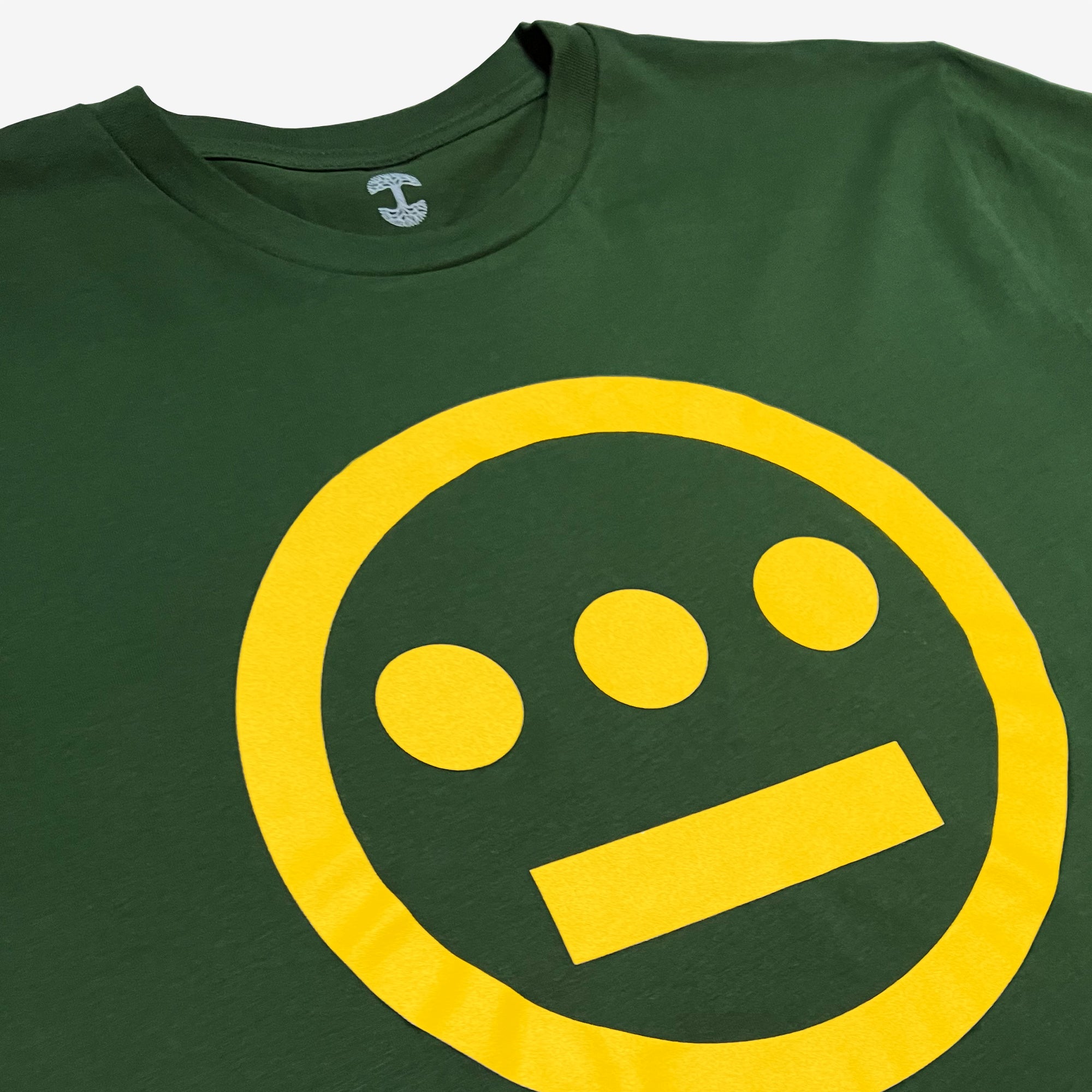 Introducing the Hiero Classic Logo Tee by Hieroglyphics, a classic fit green T-shirt showcasing a bold yellow minimalist design in the center. The design features a prominent yellow circle with three small yellow dots horizontally aligned at the top and a horizontal yellow bar near the bottom, evoking imagery of hieroglyphics or a stylized face.