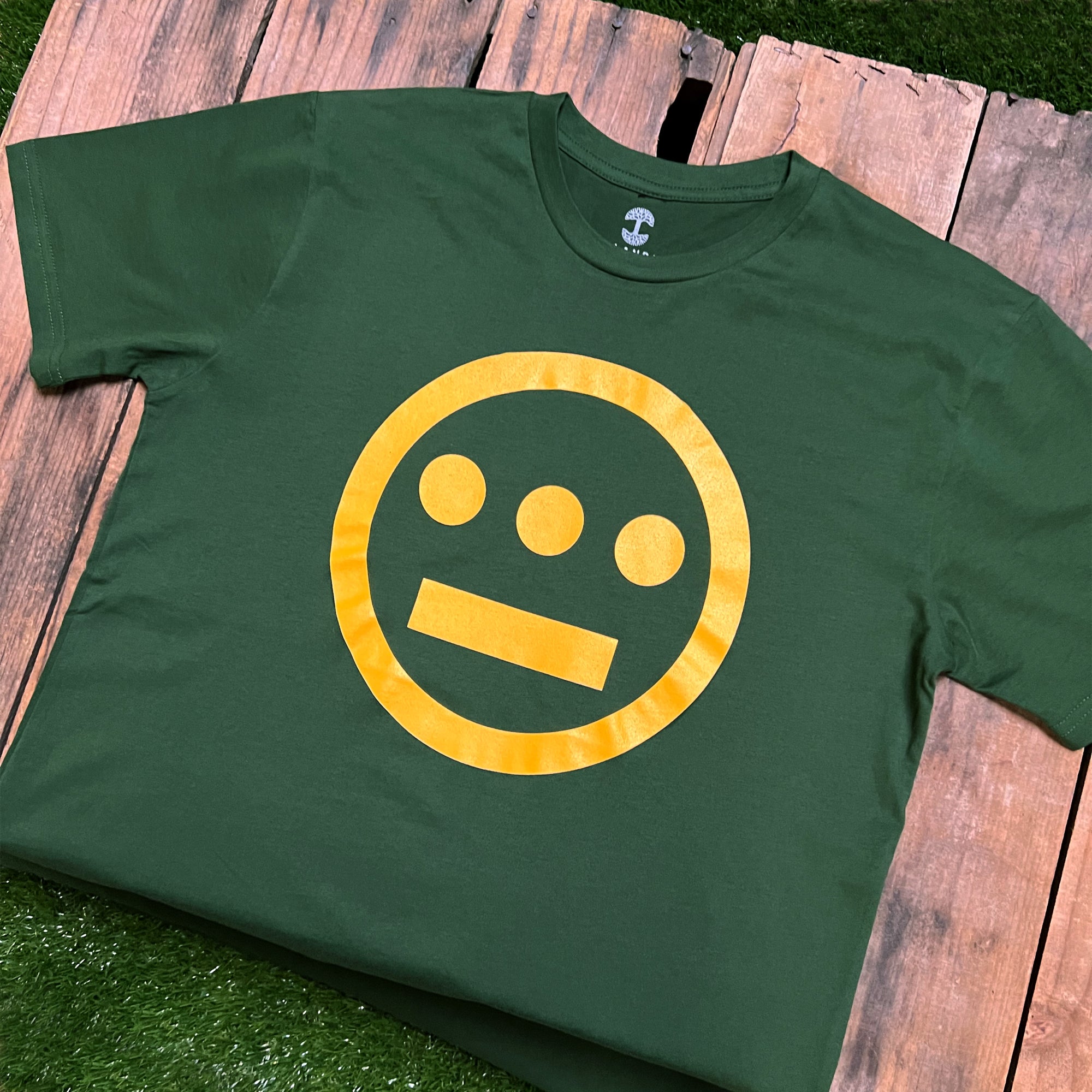 The Hiero Classic Logo Tee from Hieroglyphics, a green classic fit t-shirt, features a yellow symbol reminiscent of hieroglyphics: a circle with three dots arranged horizontally above a straight line, resembling a face. The shirt is laid flat on a wooden surface with green grass partially visible underneath.
