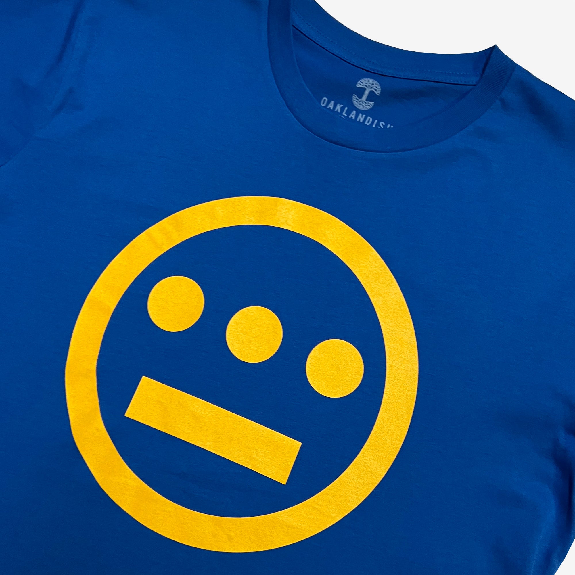 A close-up of the Women's Hiero Classic Logo Tee in royal blue, featuring a bold, yellow graphic on the chest. The graphic consists of a yellow circle containing three dots aligned horizontally above a straight line, resembling hieroglyphics. The inside tag is visible, displaying "OAKLANDISH" in white text on a darker blue background.