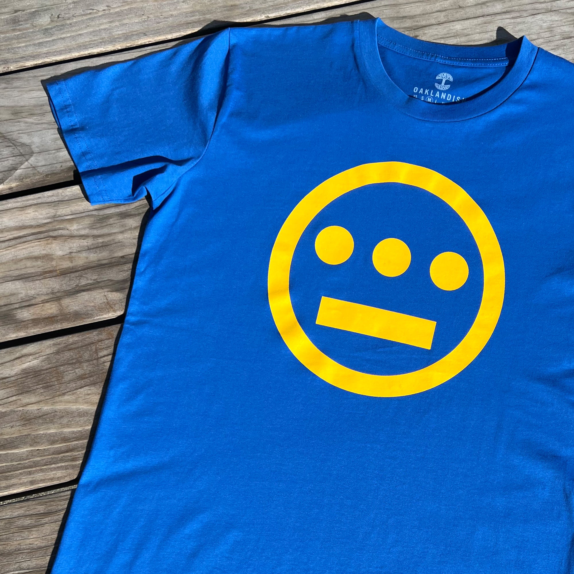 A blue Hiero Classic Logo Tee from Hieroglyphics lies flat on a wooden surface. The shirt features a yellow, circular logo with three dots in a line at the top and a horizontal line below, resembling a hieroglyphics-inspired face. The collar has text that reads "OAKLANDISH.