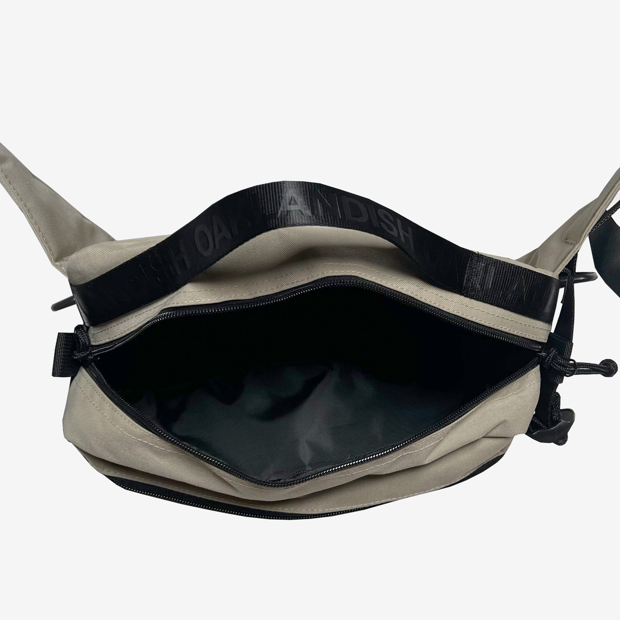 The Oaklandish Hip Bag is a beige crossbody featuring an open top view that reveals a spacious black inner compartment. The black strap has embossed text that partially reads "OAH AN DISH." This hip/shoulder bag from the Oaklandish brand showcases a zippered closure and minimalist design, set against a plain white background.