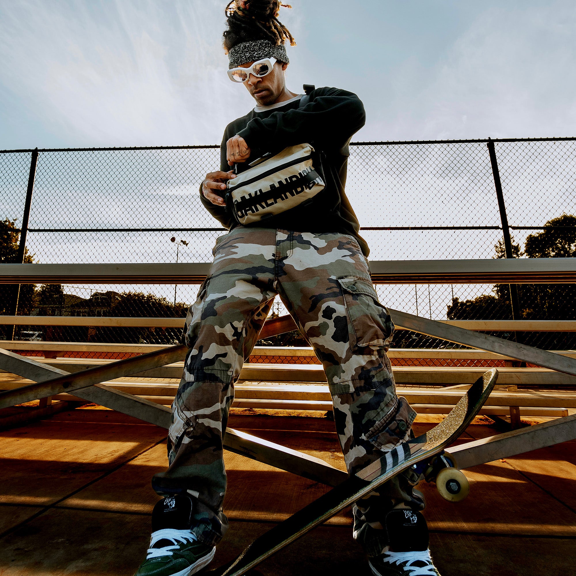 A skateboarder with dreadlocks, wearing a black sweatshirt, camo pants, white sunglasses, and a headband, holds an Oaklandish Hip Bag. They stand on bleachers next to a skateboard. The background features a chain-link fence and trees under a clear blue sky.