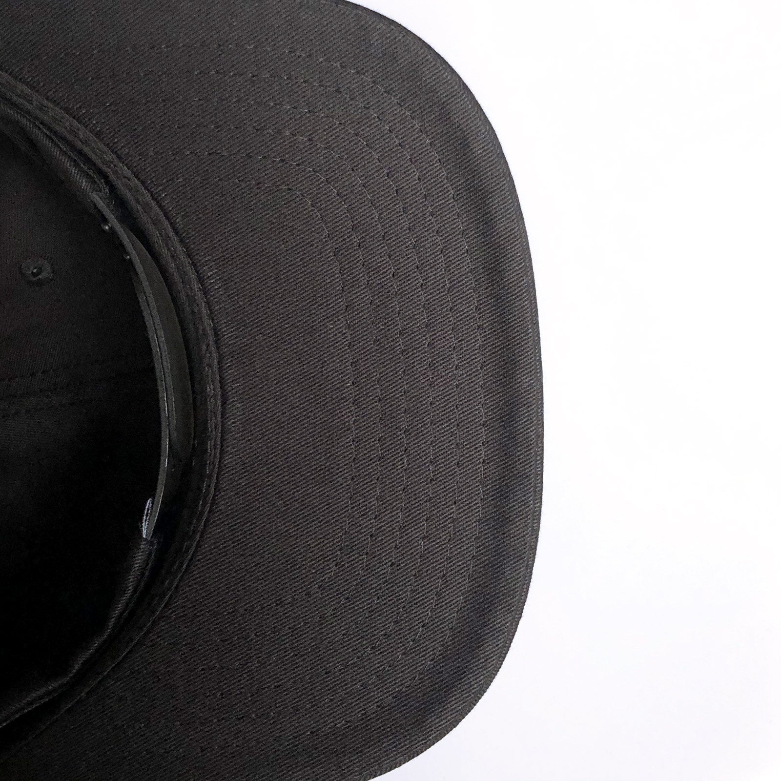Close-up image of the underside of a Greenback Snapback's brim. The cap's fabric texture and stitched lines are clearly visible. The image focuses on the front part of the brim with a white background on the right side. This headwear style, from Oaklandish, combines both comfort and urban flair.