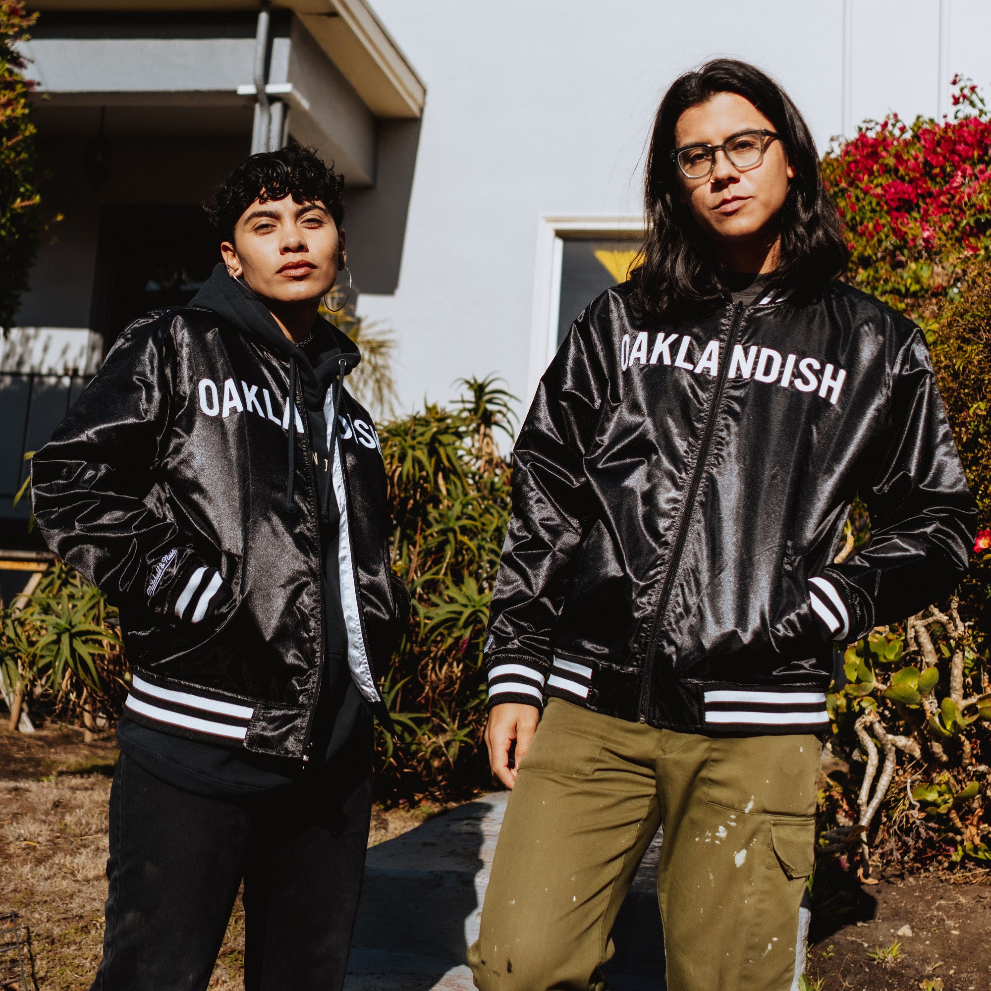 Two individuals pose confidently outdoors, against greenery and a building: one with short curly hair wears a black Oaklandish jacket over a hoodie, while the other with long straight hair pairs olive green pants with an Other Mitchell & Ness x Oaklandish Reversible Satin Jacket.