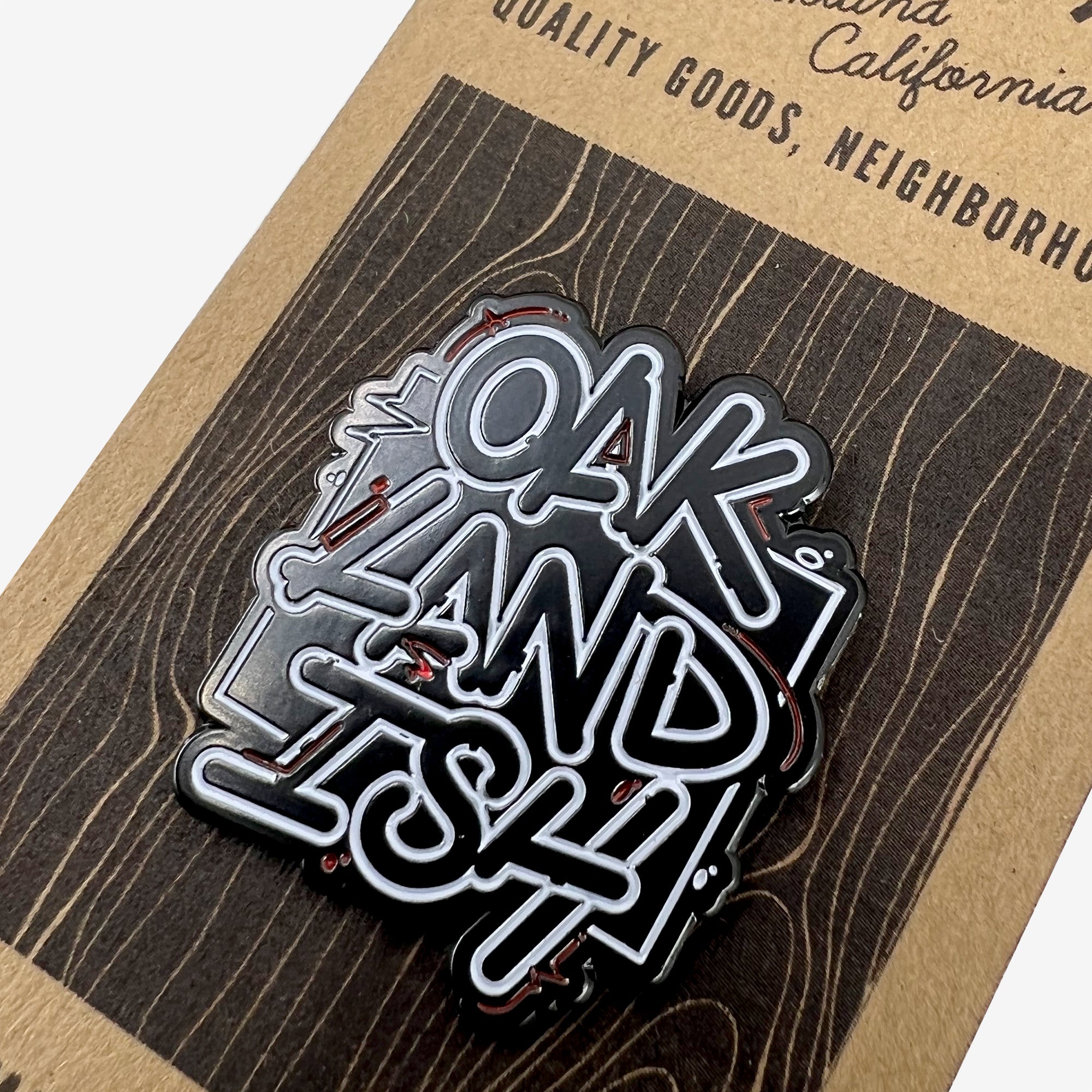 The Enamel Pin - Movement by Oaklandish features the stylized text "OAKLANDISH" in white letters on a black enamel background. Displayed on a cardboard backing that imitates a wood grain texture, it also includes partial text reading "QUALITY GOODS, NEIGHBORH...". This pin is perfect for accessorizing any outfit.