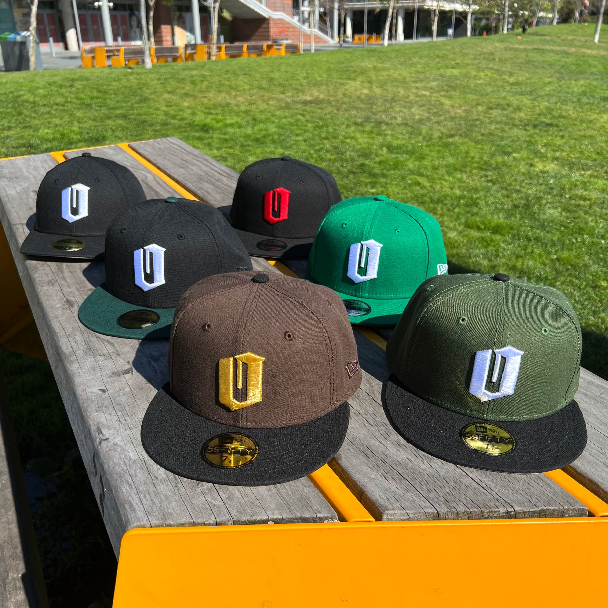 Seven New Era Official 59FIFTY Fitted Caps from Oaklandish are displayed on a wooden bench outdoors. The caps come in various colors, including black, green, brown, and dark green, each featuring a large embroidered "U" on the front in different hues like white, gold, and red. A grassy area and some trees can be seen in the background.