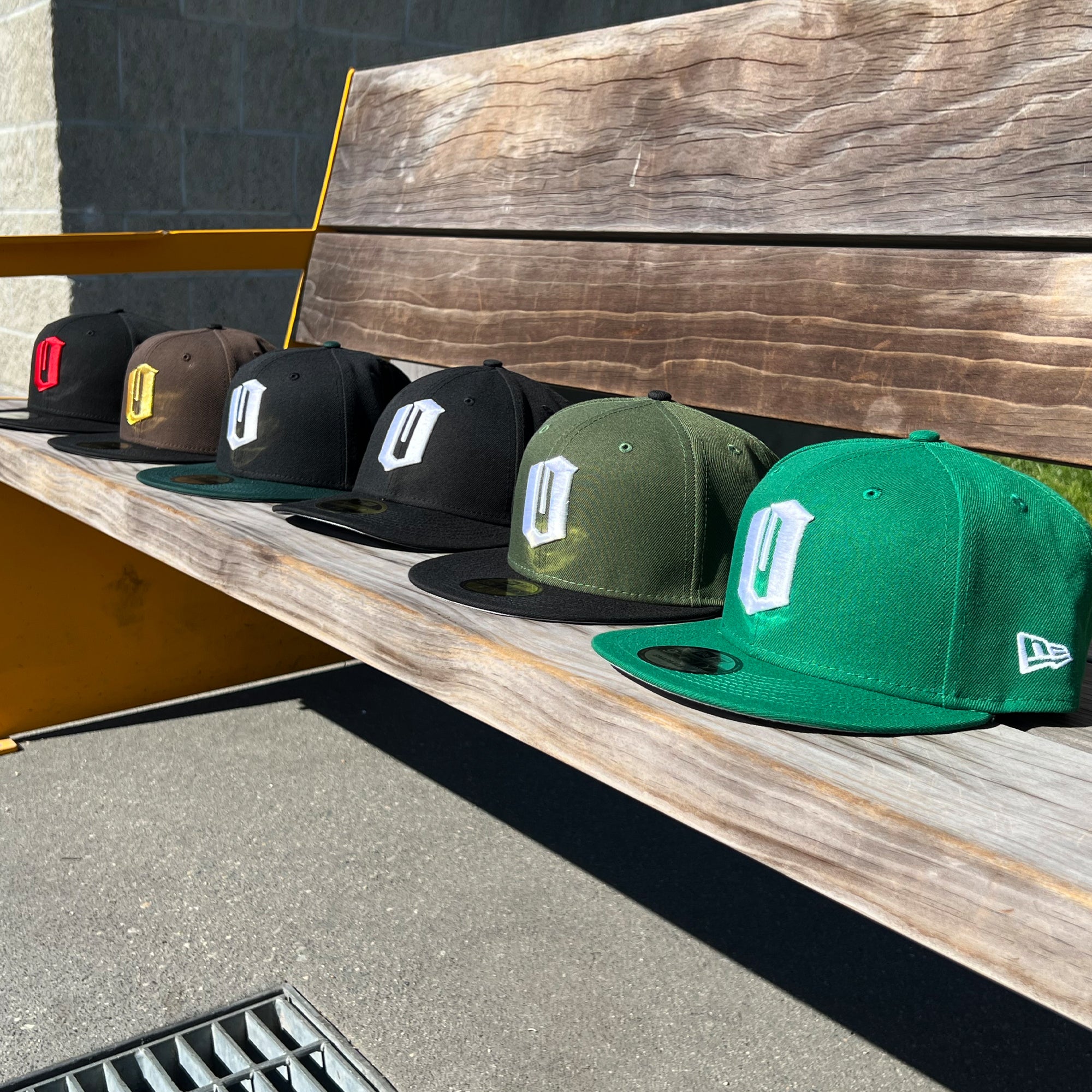 Six New Era Official 59FIFTY Fitted Caps from Oaklandish are neatly arranged on a wooden bench outdoors. Available in an array of colors including black, brown, dark green, olive green, and bright green, each cap showcases a monogrammed "D" on the front and the iconic Oaklandish logo on the side.