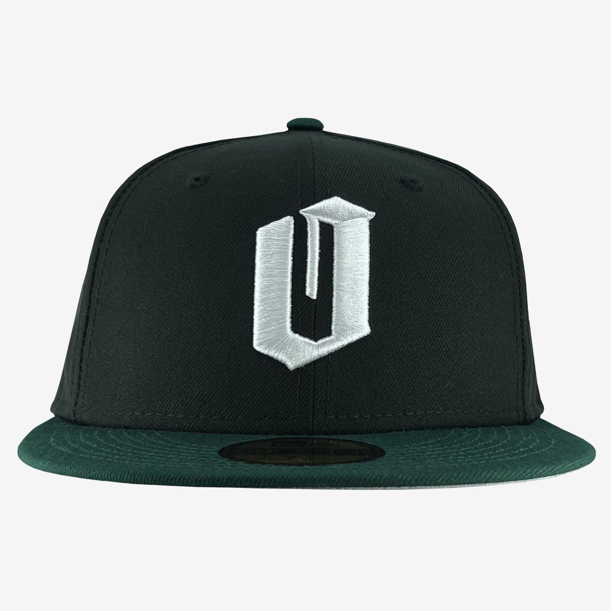 The Oaklandish New Era Official 59FIFTY Fitted Cap is a black hat with a dark green flat brim. The front showcases a large, bold, white embroidered letter "U" in a stylized font. The button on top of the cap matches the main black color.