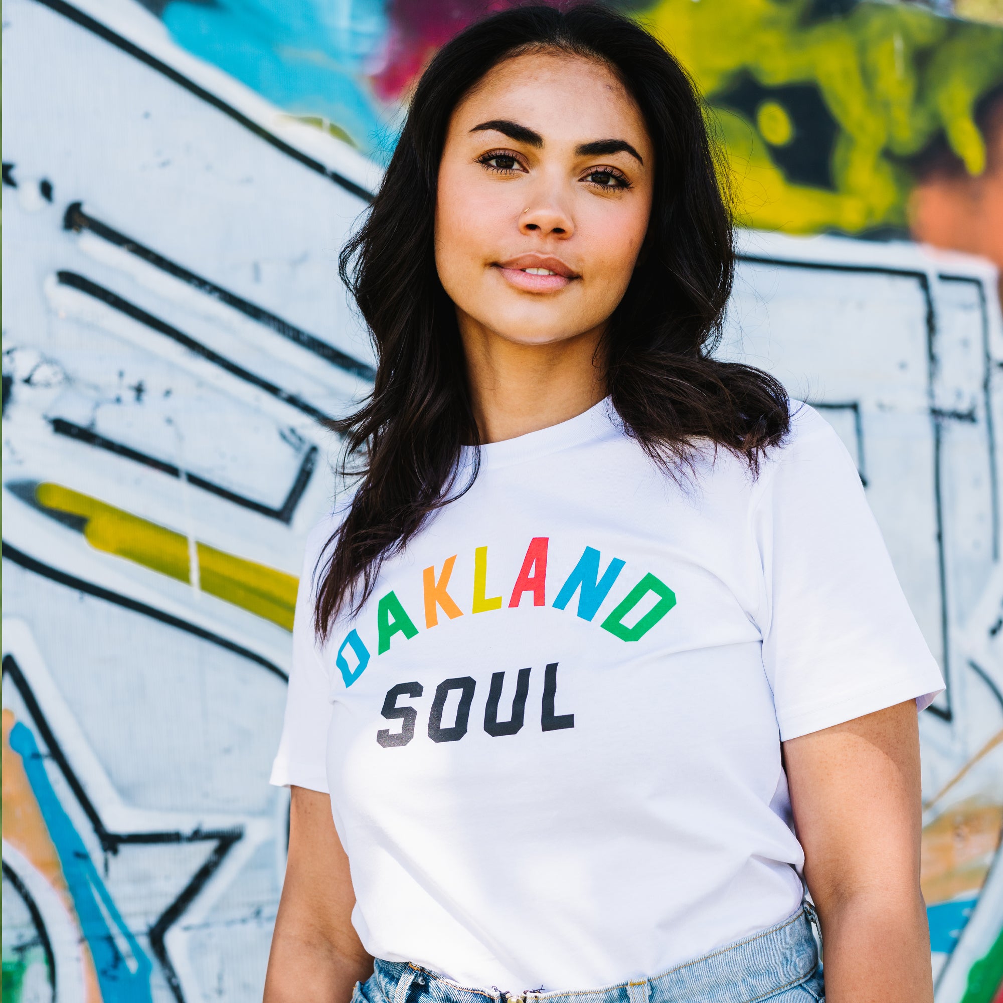 A woman with long dark hair stands in front of a colorful graffiti wall. She wears an Oakland Roots SC Oakland Soul Wordmark Tee, featuring "OAKLAND SOUL" in rainbow-colored and black letters. Gazing confidently at the camera with a slight smile, her hands are tucked into the pockets of her light-blue jeans.