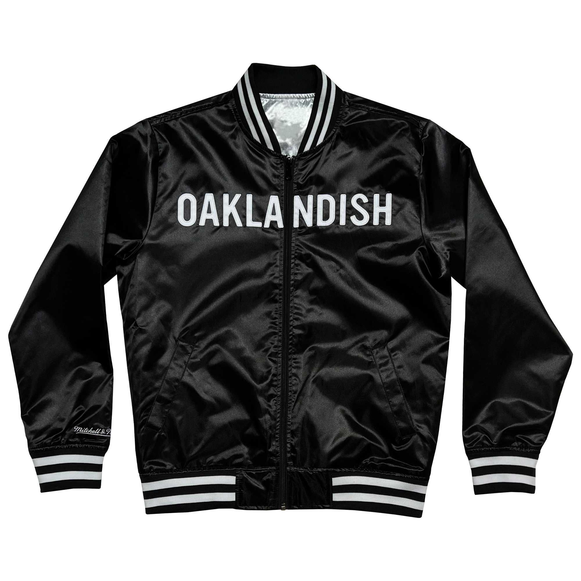 A black reversible satin jacket from the Mitchell & Ness x Oaklandish collaboration features bold white "OAKLANDISH" across the chest, striped collar, cuffs, waistband, and a signature on the bottom left front, elegantly displayed against a white background.