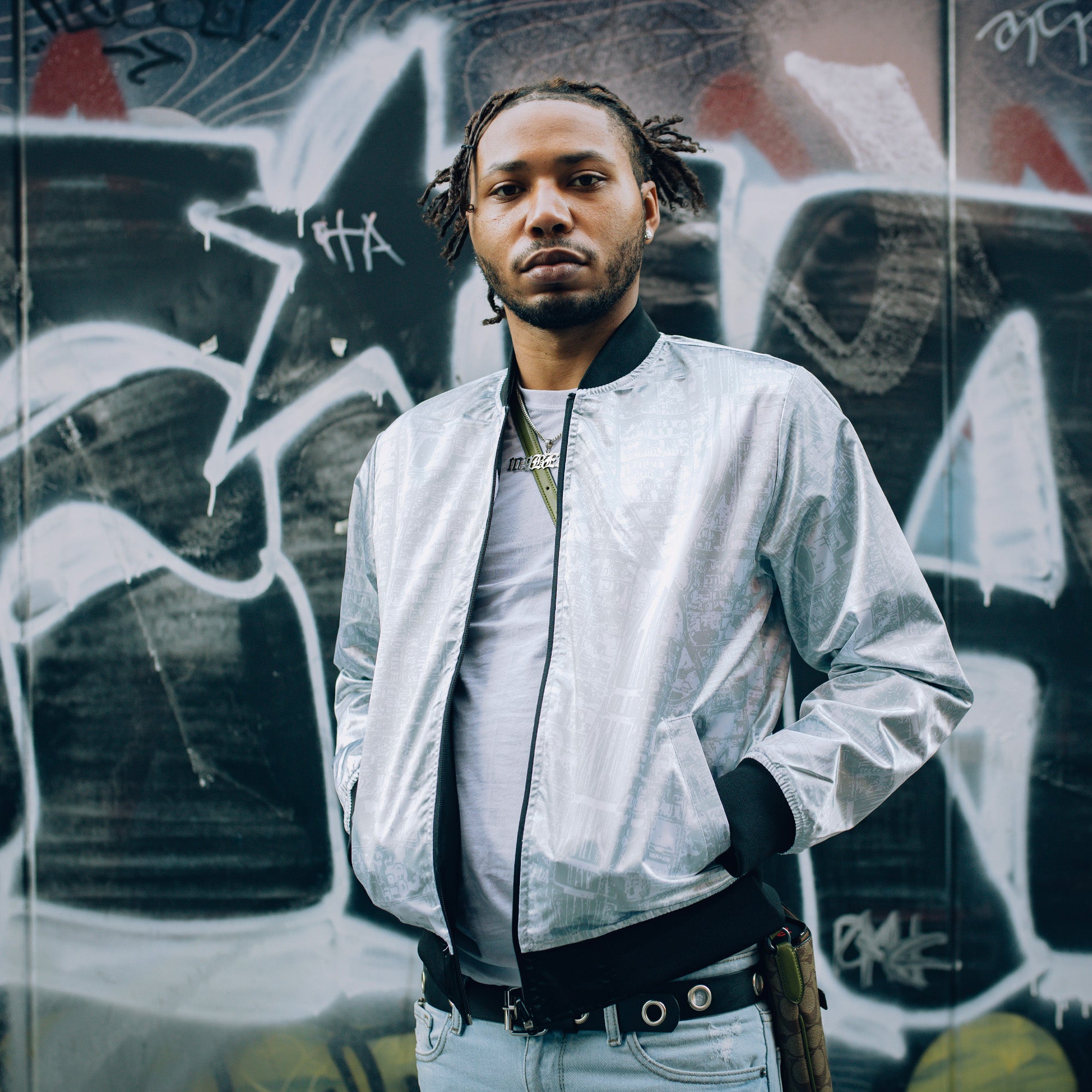 Wearing a silver Mitchell & Ness x Oaklandish Reversible Satin Jacket, a person with short dreadlocks poses confidently next to a graffiti-covered wall. Their jeans have a chain at the waist, and the backdrop showcases abstract patterns in black, white, and gray tones.