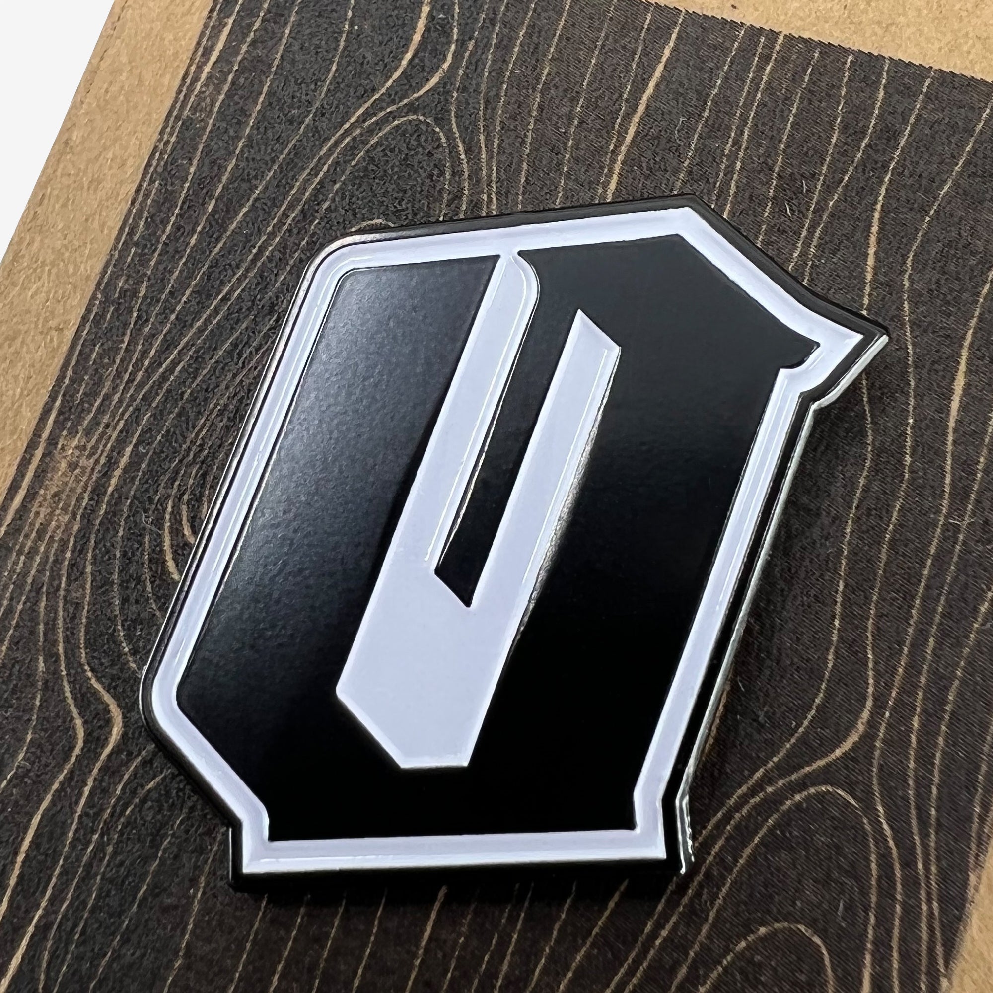 Close-up image of the Oaklandish Enamel Pin - Official, featuring a metallic emblem shaped like the letter "O" with a sleek black and silver design. The enamel lapel pin is placed on a surface with a dark wood grain pattern, highlighting the contrast between the accessory's colors and the textured background.