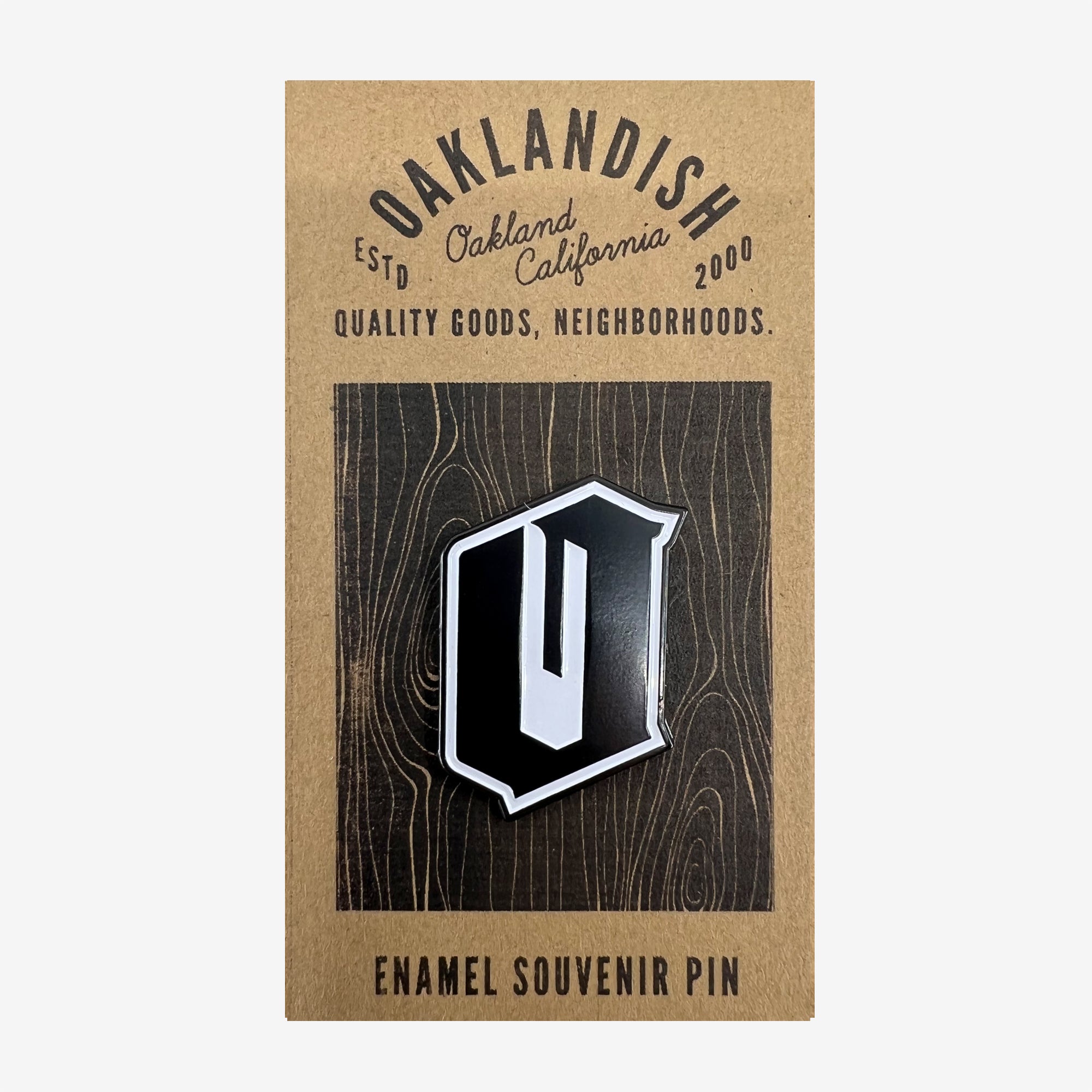 A black enamel lapel pin featuring a silver-bordered, stylized letter "O" displayed on a wooden background card. The card reads "Oaklandish, Estd Oakland California 2000, Quality Goods, Neighborhoods," and ends with "Enamel Souvenir Accessories" at the bottom.