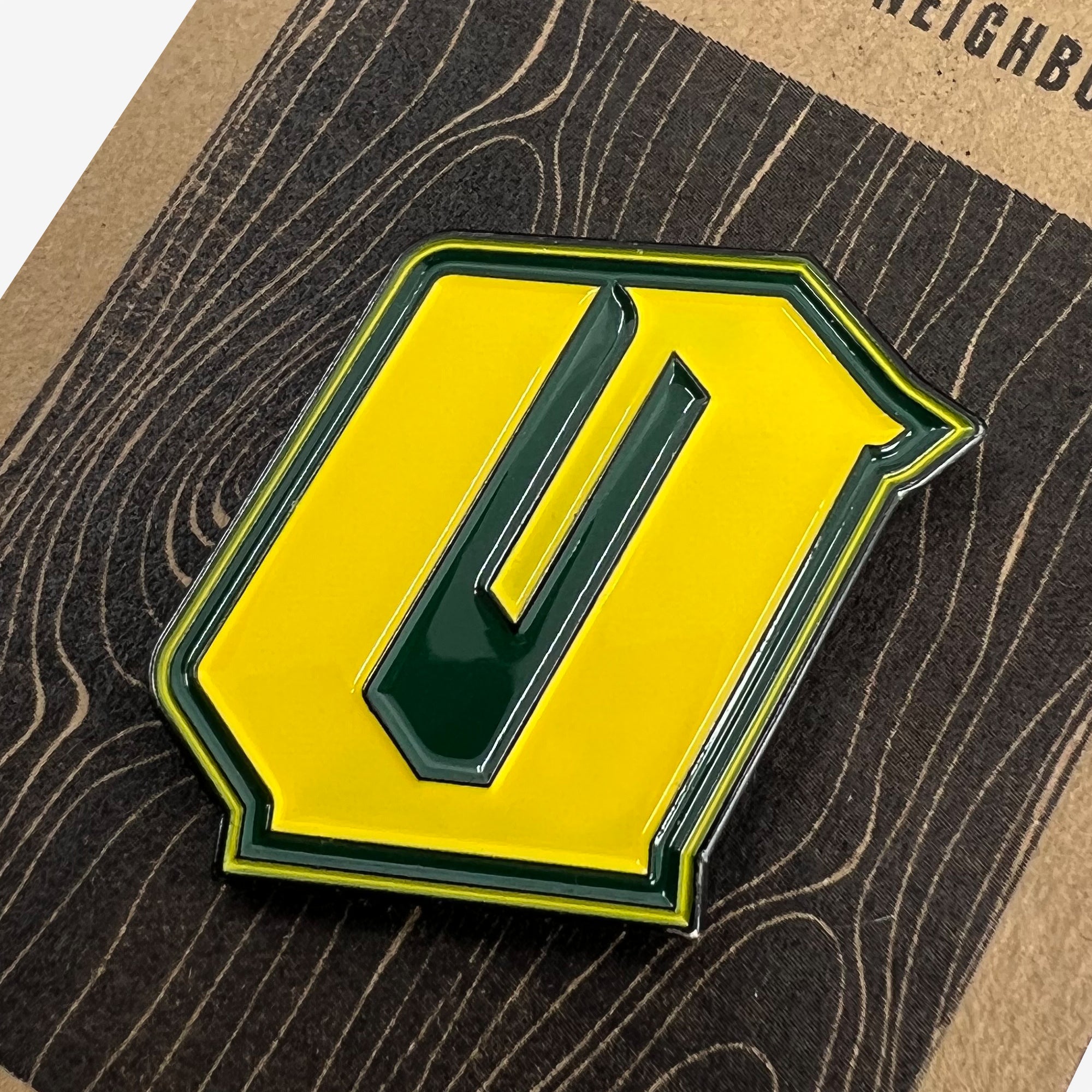 The "Enamel Pin - Official" by Oaklandish features a vibrant green letter "O" outlined in yellow, placed against a dark, textured surface. The yellow border around the green letter makes it stand out prominently. The wood grain pattern of the background adds a rustic feel to this Oakland-inspired accessory.