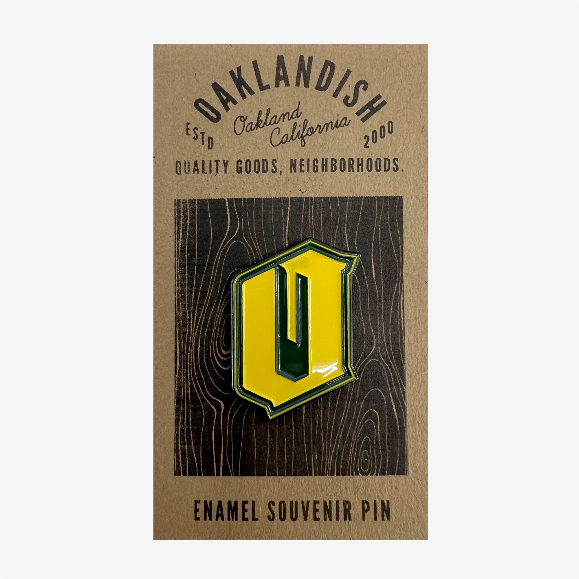 A rectangular card features the text "OAKLANDISH Estd Oakland California 2000. Quality Goods, Neighborhoods." Below the text is the Enamel Pin - Official from Oaklandish, shaped like a stylized letter 'O' in green and yellow. The bottom of the card reads "Enamel Souvenir Pin." The background has a wood grain design, perfect for highlighting your Oakland accessories.