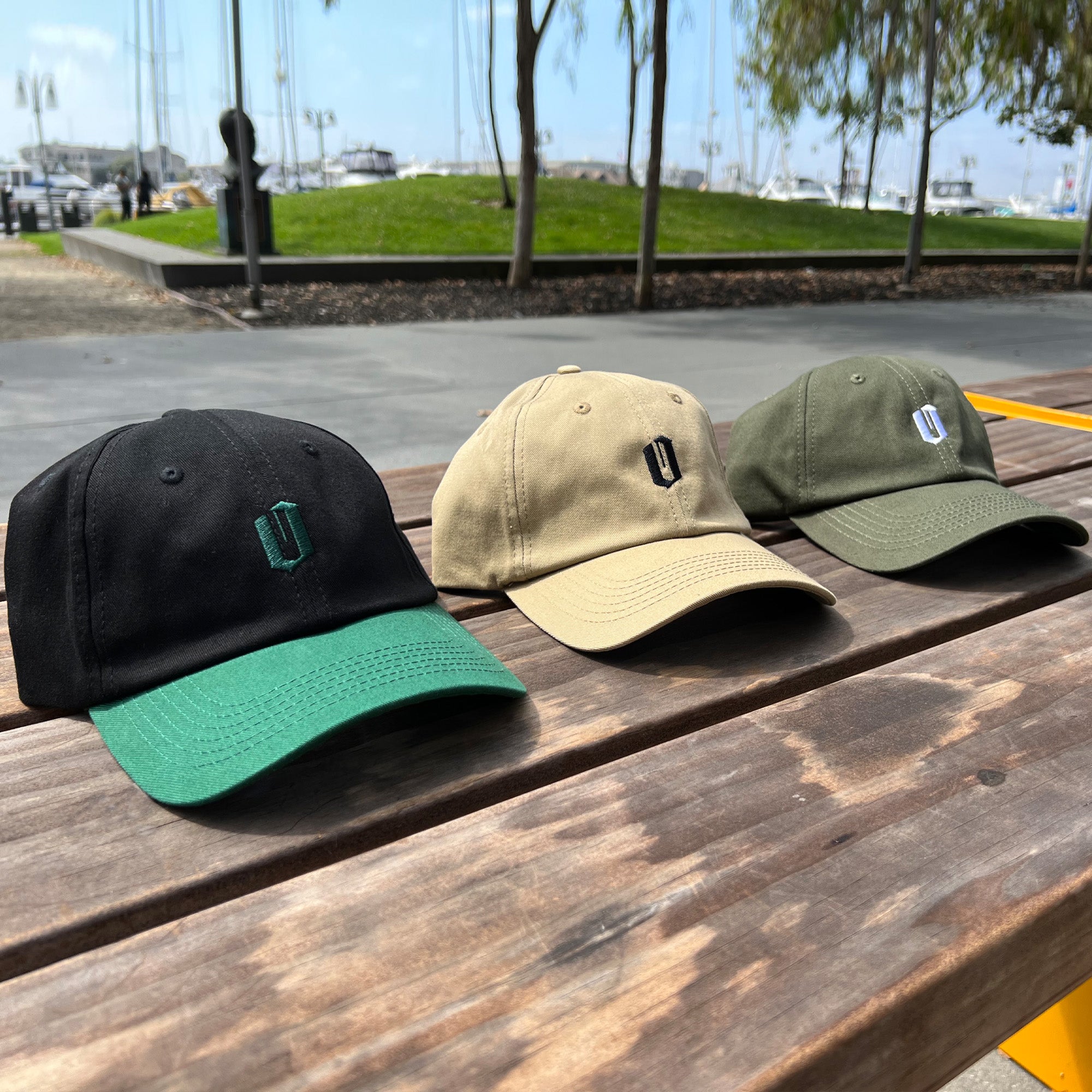 Three Official Micro Logo Dad Hats by Oaklandish displayed on a wooden bench. The caps are available in black with a green brim, beige, and olive green colors. Each hat features an embroidered "U" logo on the front and has an adjustable strap at the back. In the background, there's a marina, grassy area, and trees under the sunny skies.