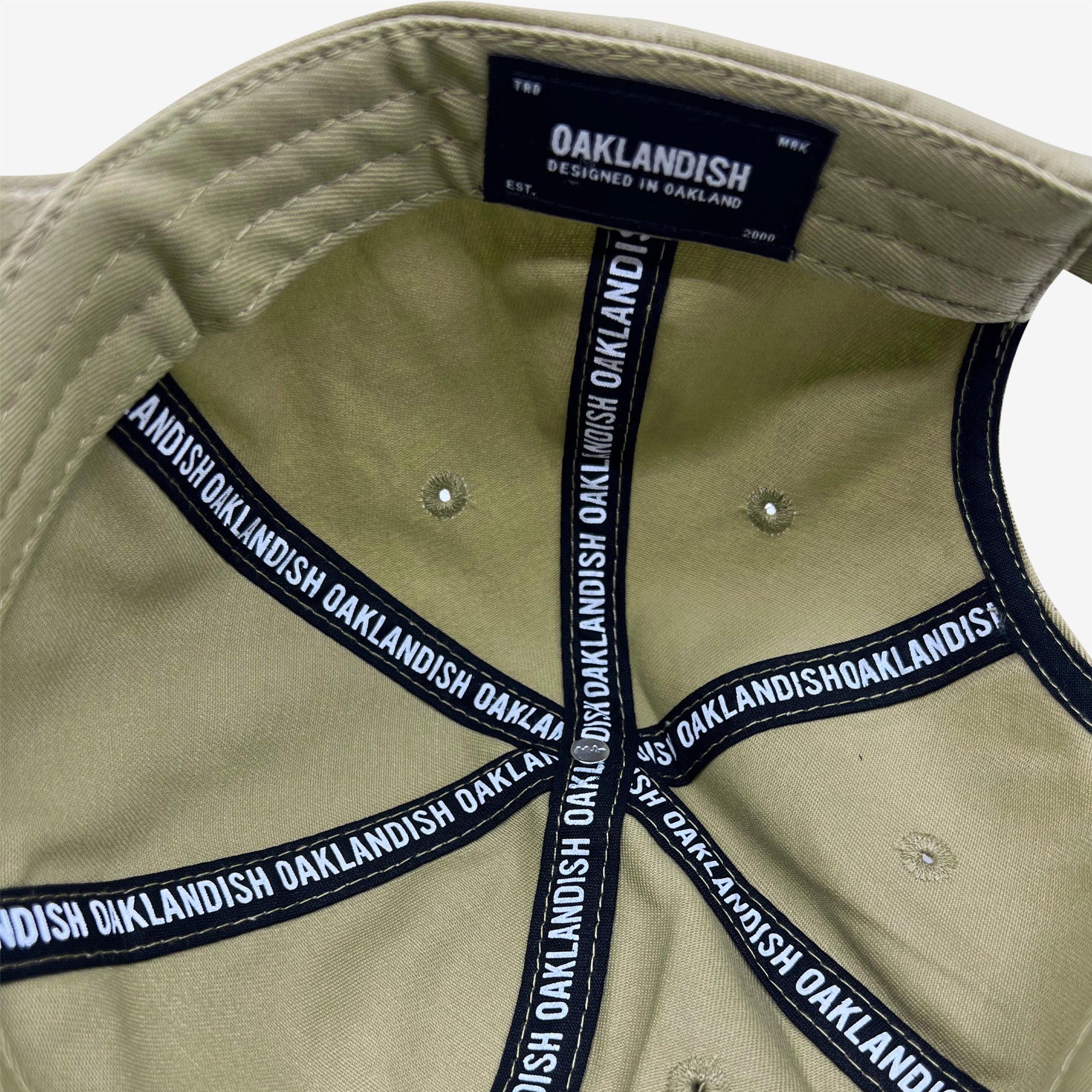 The image reveals the interior of an Oaklandish Official Micro Logo Dad Hat. The black fabric bands, imprinted with "Oaklandish" in white, fan out from the center of the crown. Inside, a tag reads, "Oaklandish, designed in Oakland, Est. 2000." The hat features an adjustable strap for a perfect fit.