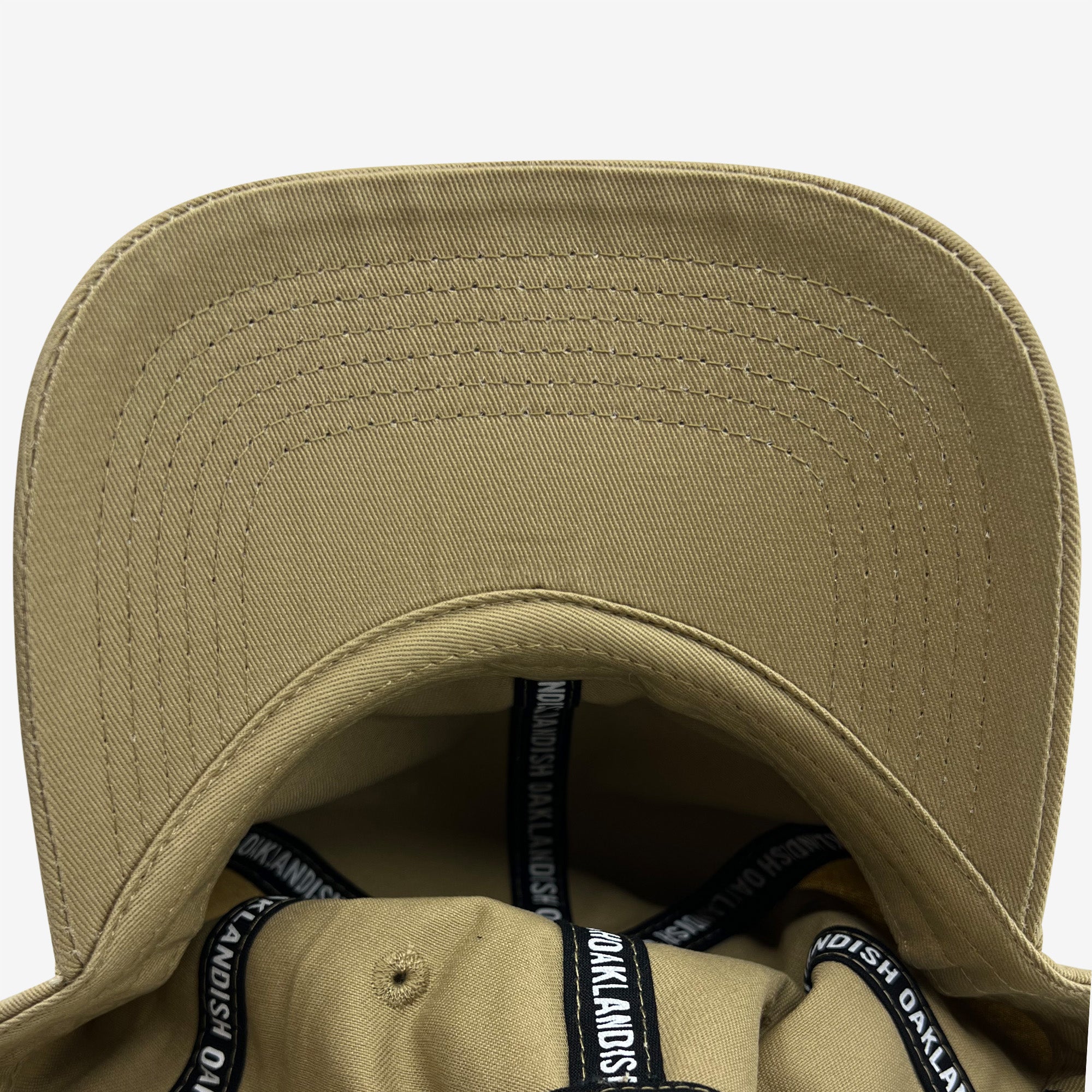 A close-up view of the inside of the Official Micro Logo Dad Hat by Oaklandish, showcasing its beige color, curved brim, and precise stitching. The inner lining is adorned with black bands featuring white text that repeatedly spells "Oaklandish" around the edges. The sturdy fabric and clear stitching are evident throughout, along with an adjustable strap for a perfect fit.