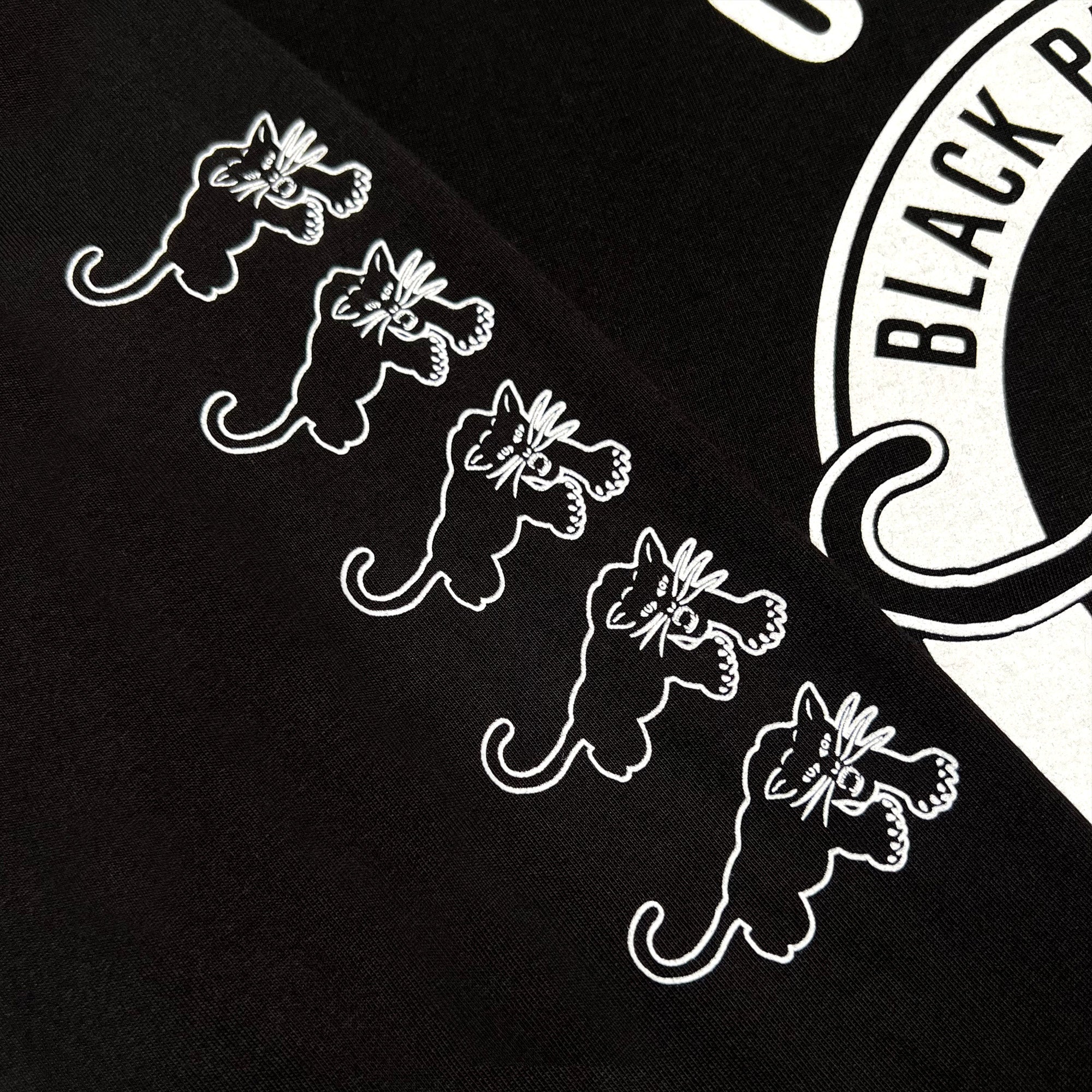 The image features the Black Panther Power LS Tee by Oaklandish, showcasing a black fabric adorned with five identical white-printed cat illustrations aligned horizontally. The cats exhibit playful, wide-eyed expressions and raised paws. To the right, there's a partial view of an ornate white circular logo, reflecting the bold legacy of Black Panther Power.