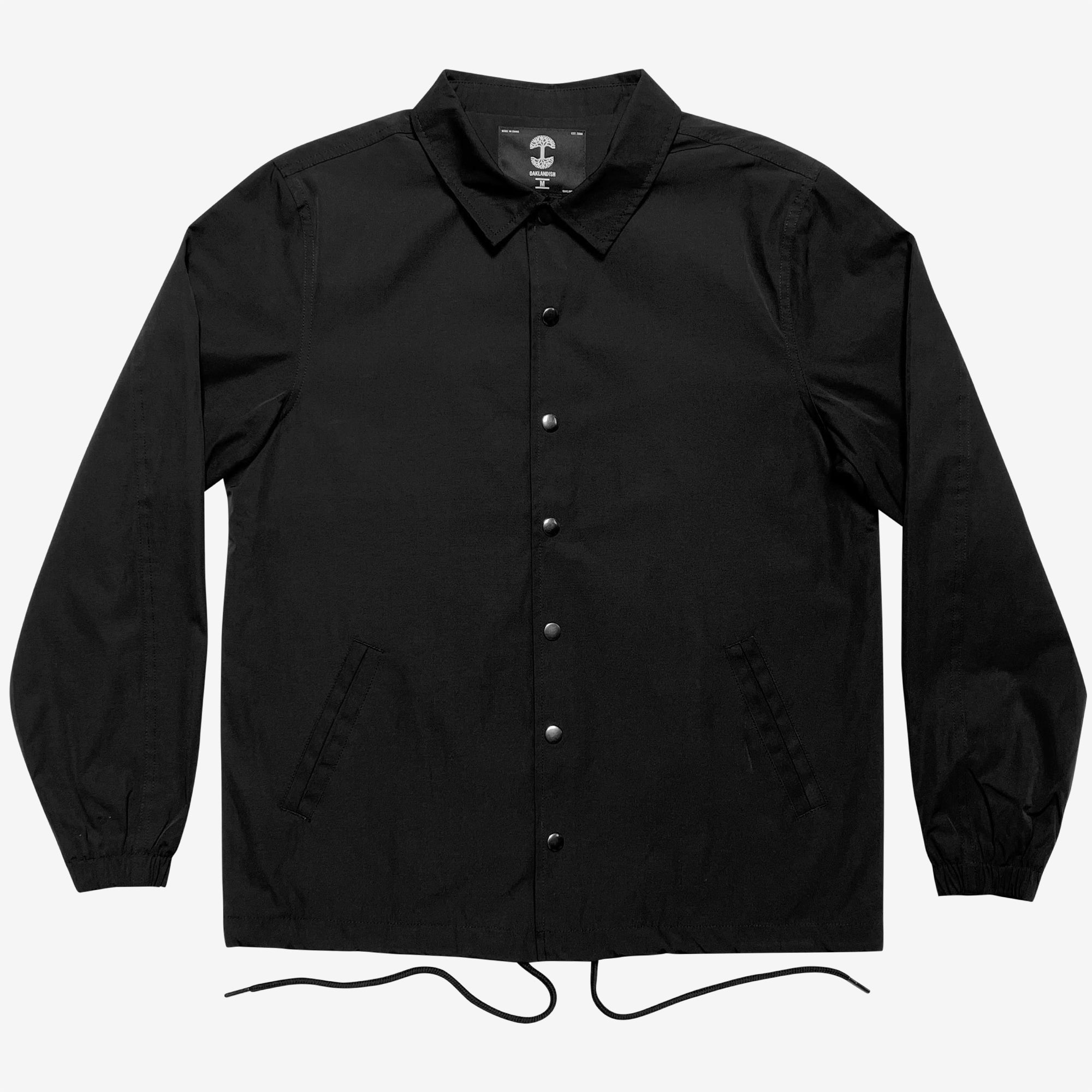 A black, front-button Premium Coaches Jacket from Oaklandish with a pointed collar is displayed on a plain white background. The water-resistant jacket features long sleeves with elastic cuffs, two slanted side pockets, and an adjustable drawstring at the hem. Designed in men's sizing, it has a tag with white graphics and text at the inside back collar.