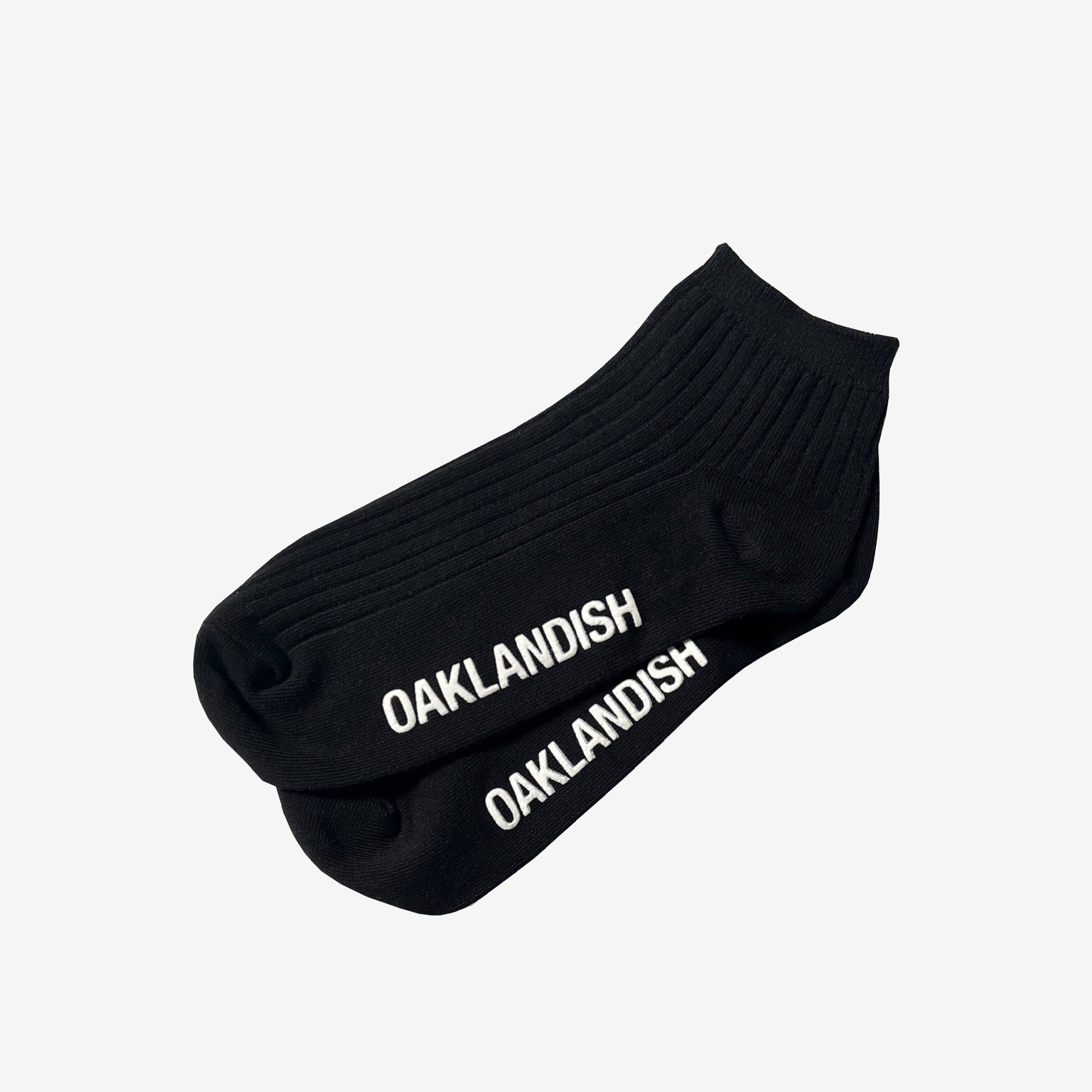 A pair of Grip Low Crew Socks from Oaklandish, featuring a black, ribbed design with "OAKLANDISH" printed in white capital letters along the top, are laid flat against a plain white background.