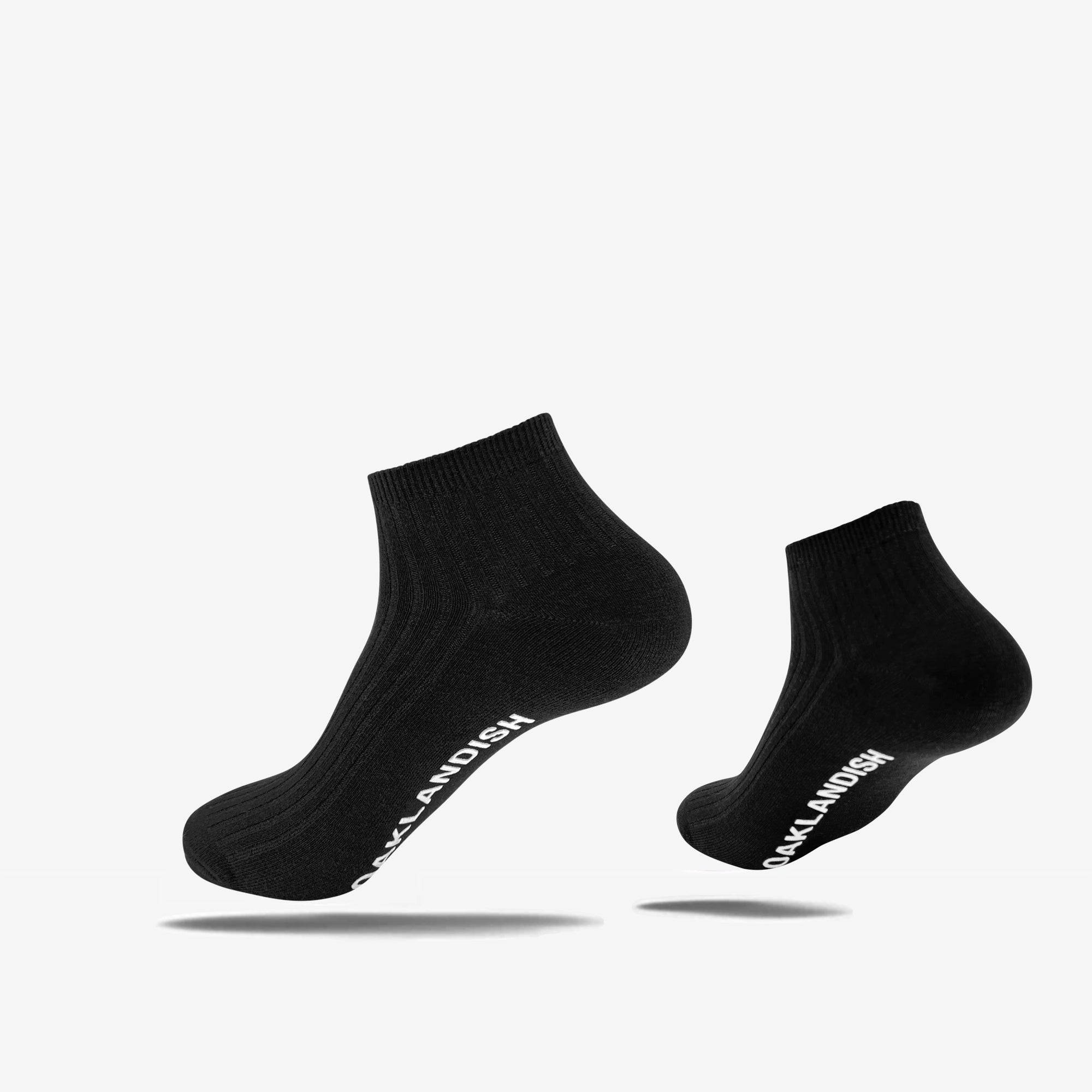 The Grip Low Crew Sock by Oaklandish is displayed against a simple white background. These black socks feature a ribbed texture and have "SOCKLANDISH" written in white text along the outside of the arch. The socks appear to be slightly floating, casting shadows on the surface below them.