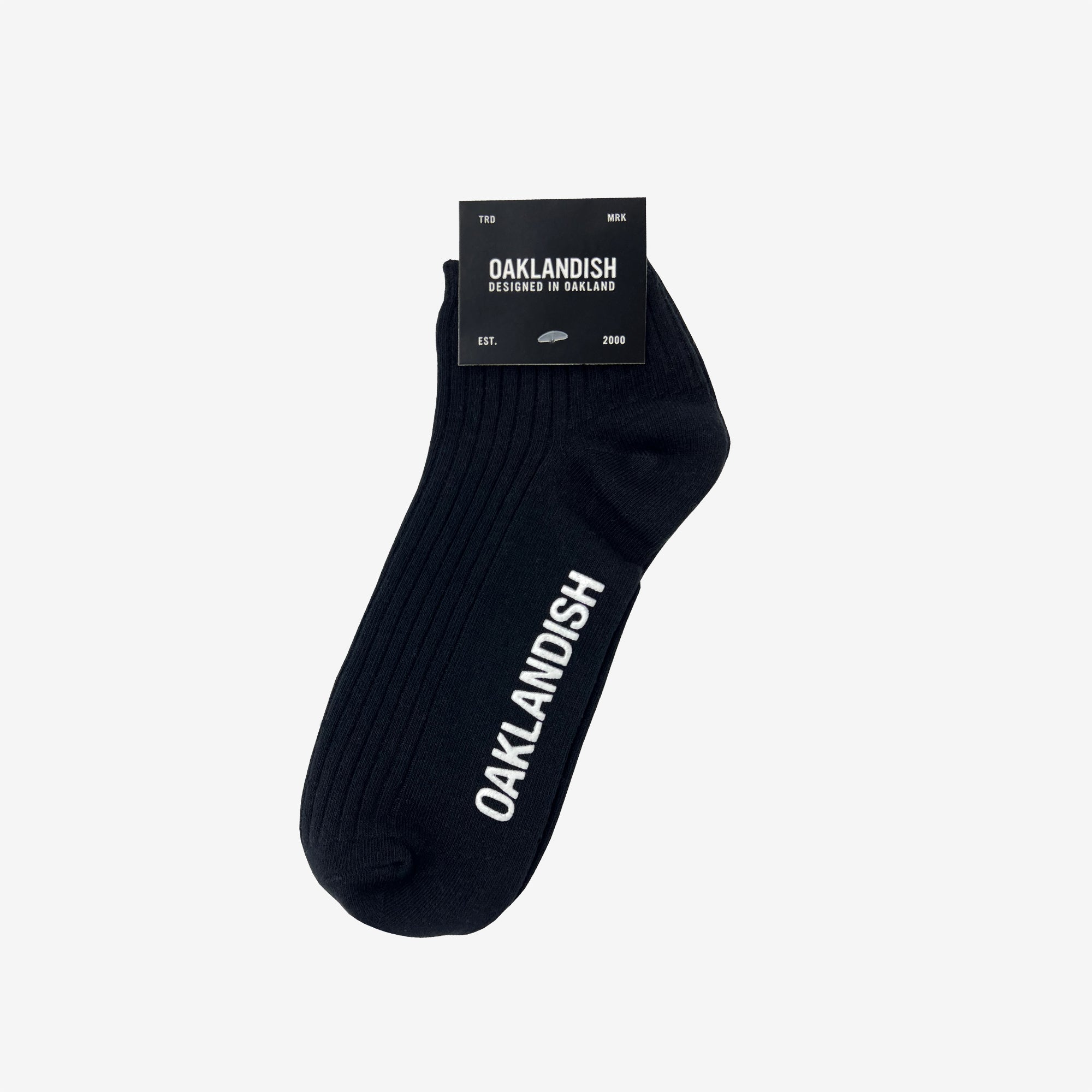 A single black "Grip Low Crew Sock" by Oaklandish features the word "OAKLANDISH" printed in white along its side. This classic example of Oaklandish crew socks comes attached to black packaging that reads "OAKLANDISH," "Designed in Oakland," "EST. 2000," and size information labeled as "TBD MK." The sock is displayed against a plain white background.